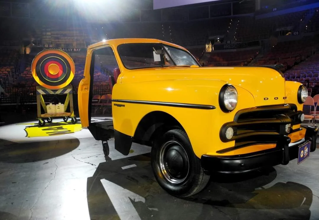 A vintage yellow taxi cab on display inside an arena with seating visible in the background, and a large colorful target prop next to it.