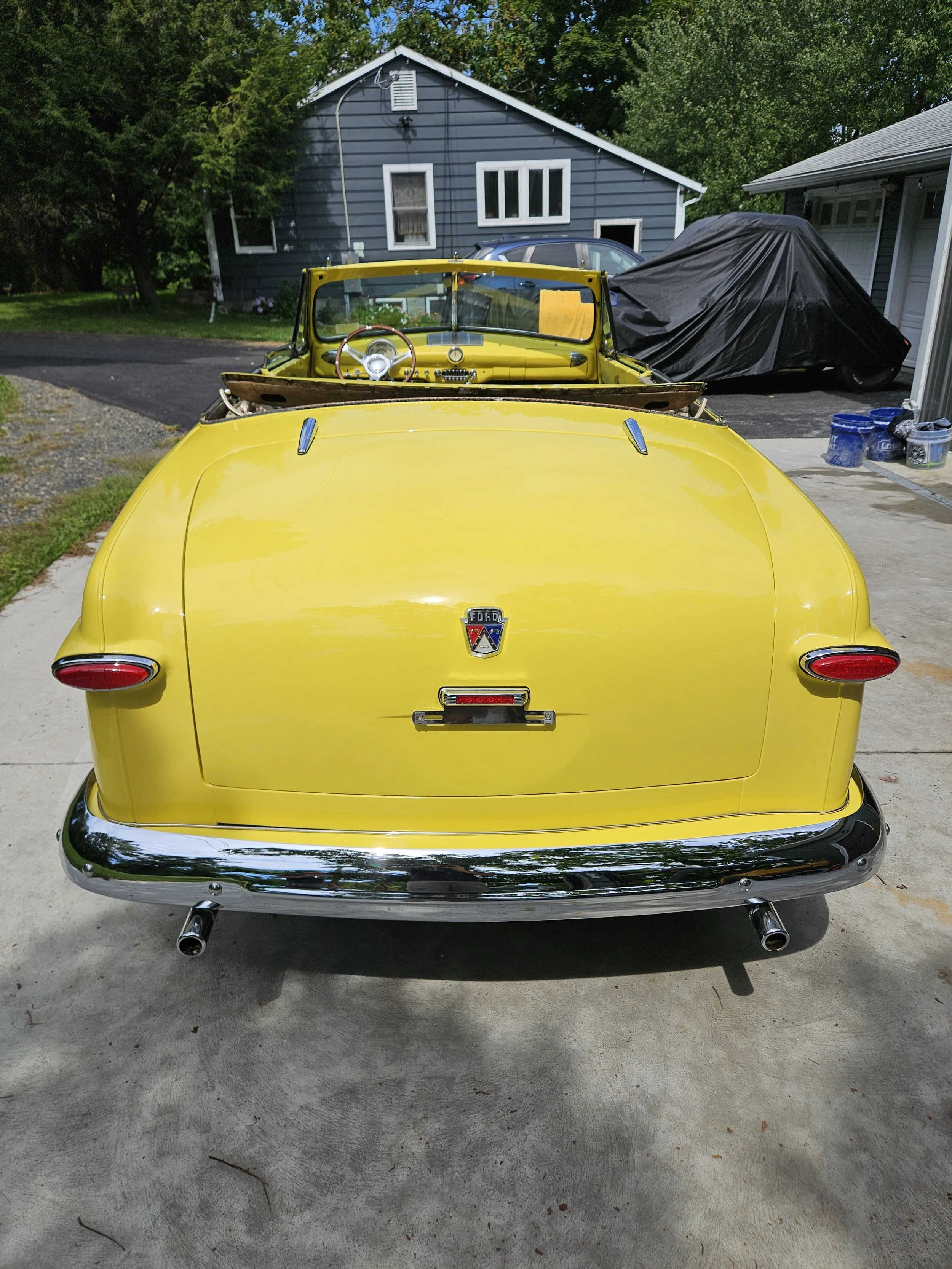 Rear view of a vintage yellow Ford convertible car parked on a concrete driveway in front of a house with a blue exterior. The car has chrome accents, dual exhaust pipes, and a Ford emblem on the trunk.