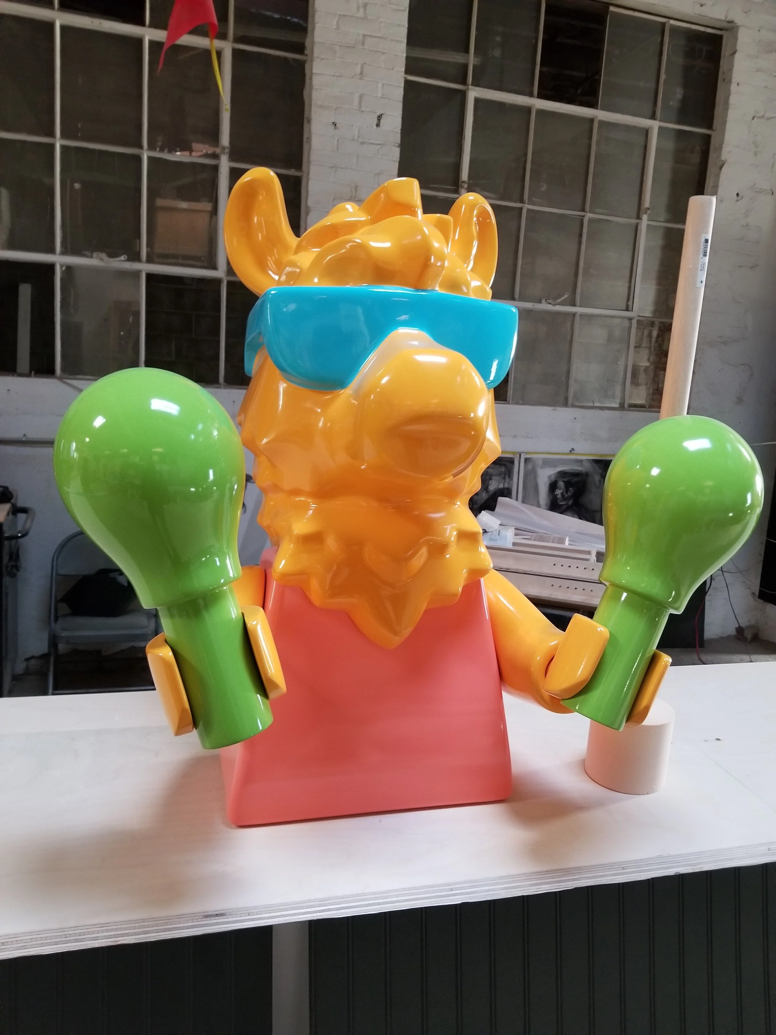 Colorful sculpture of a camel wearing blue sunglasses, with green maracas, pink shirt, set against an industrial background.