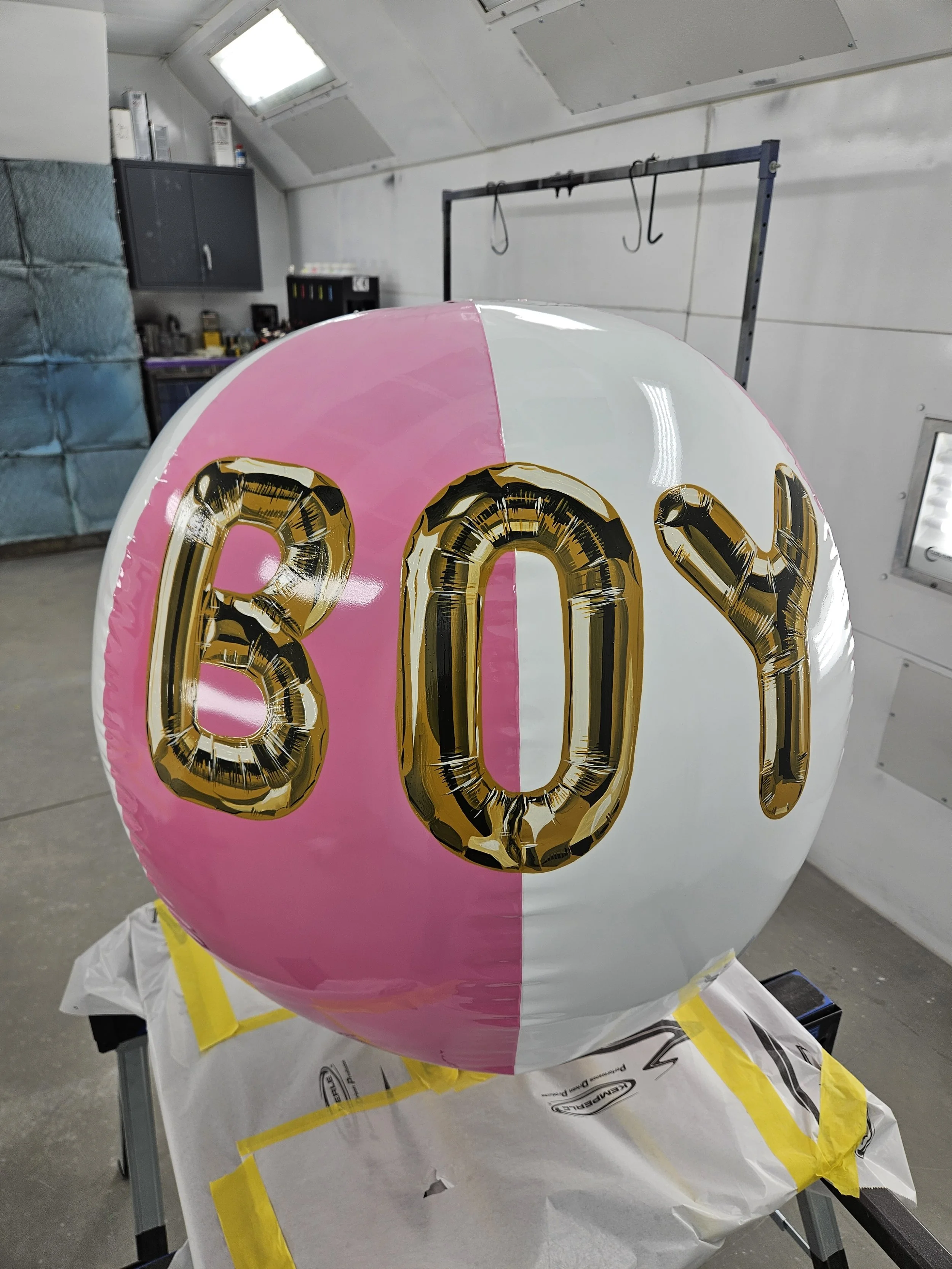 A round balloon with pink and white halves, displaying the word 'BOY' in large gold and black letters.