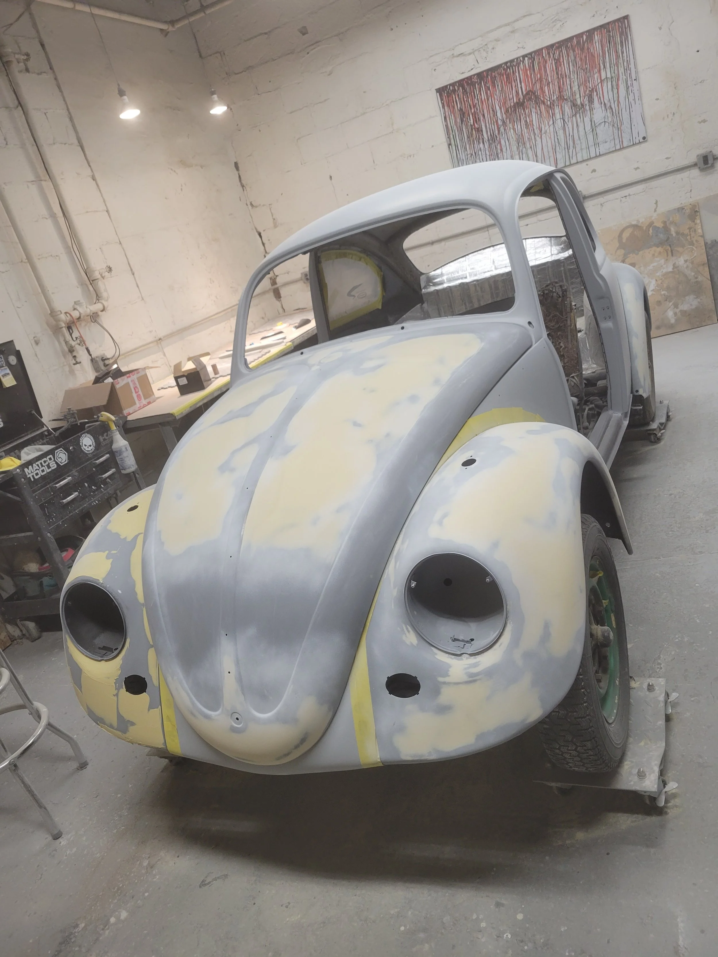 A vintage Volkswagen Beetle car under restoration, with parts and primer on its body in a workshop.