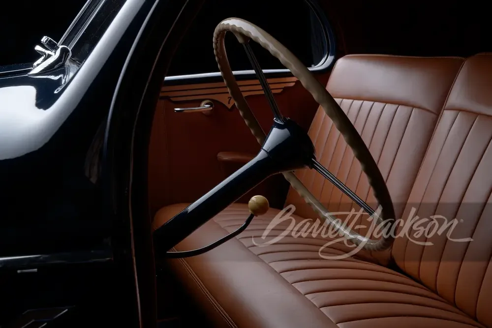 Interior of a vintage car showing a brown leather seat, a steering wheel, and a gear shift, with a black exterior window frame.
