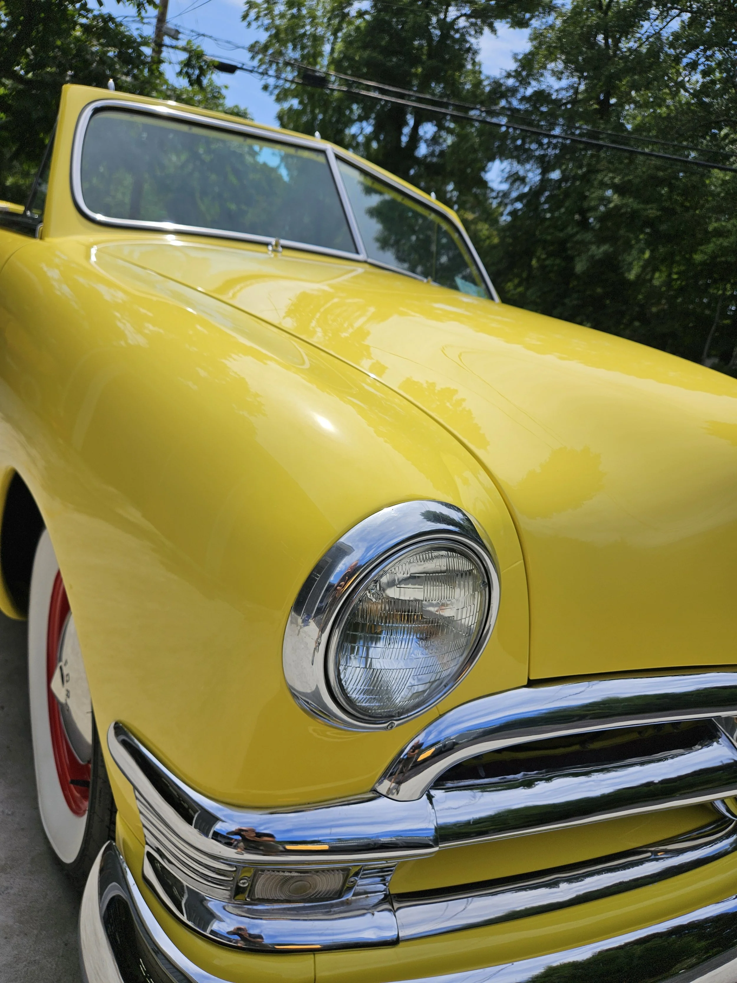Close-up of a vintage yellow car with chrome bumper and wheel arch, reflecting the surroundings, set outdoors with trees and clear sky in the background.