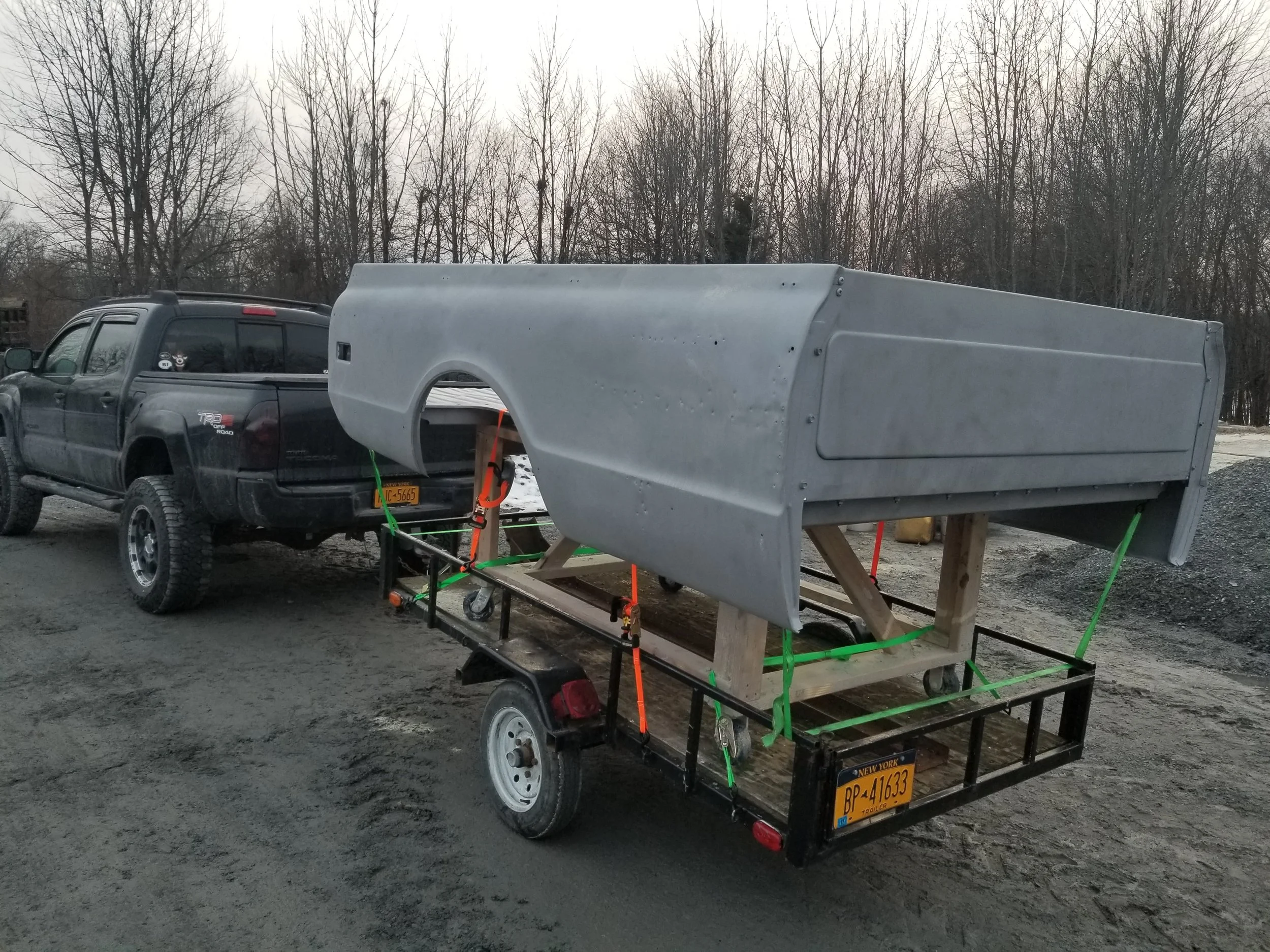 A black pickup truck is parked on a dirt road with a large, light gray boat shell secured on a wooden and metal trailer behind it.