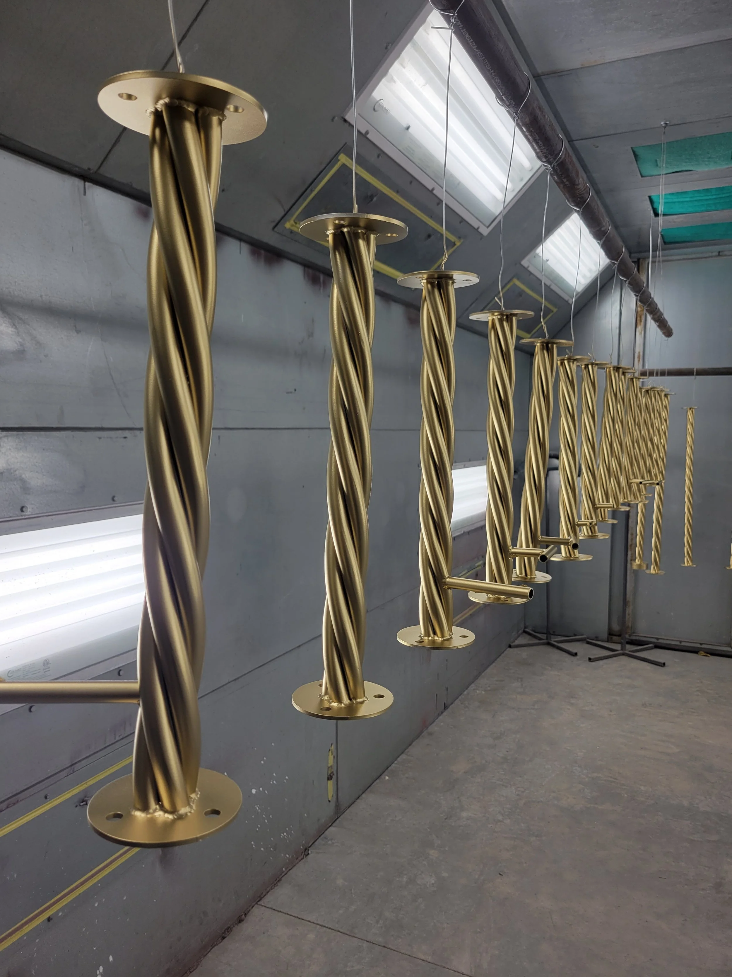 Multiple gold-colored twisted metal rods hanging from the ceiling, possibly in a workshop or manufacturing setting.