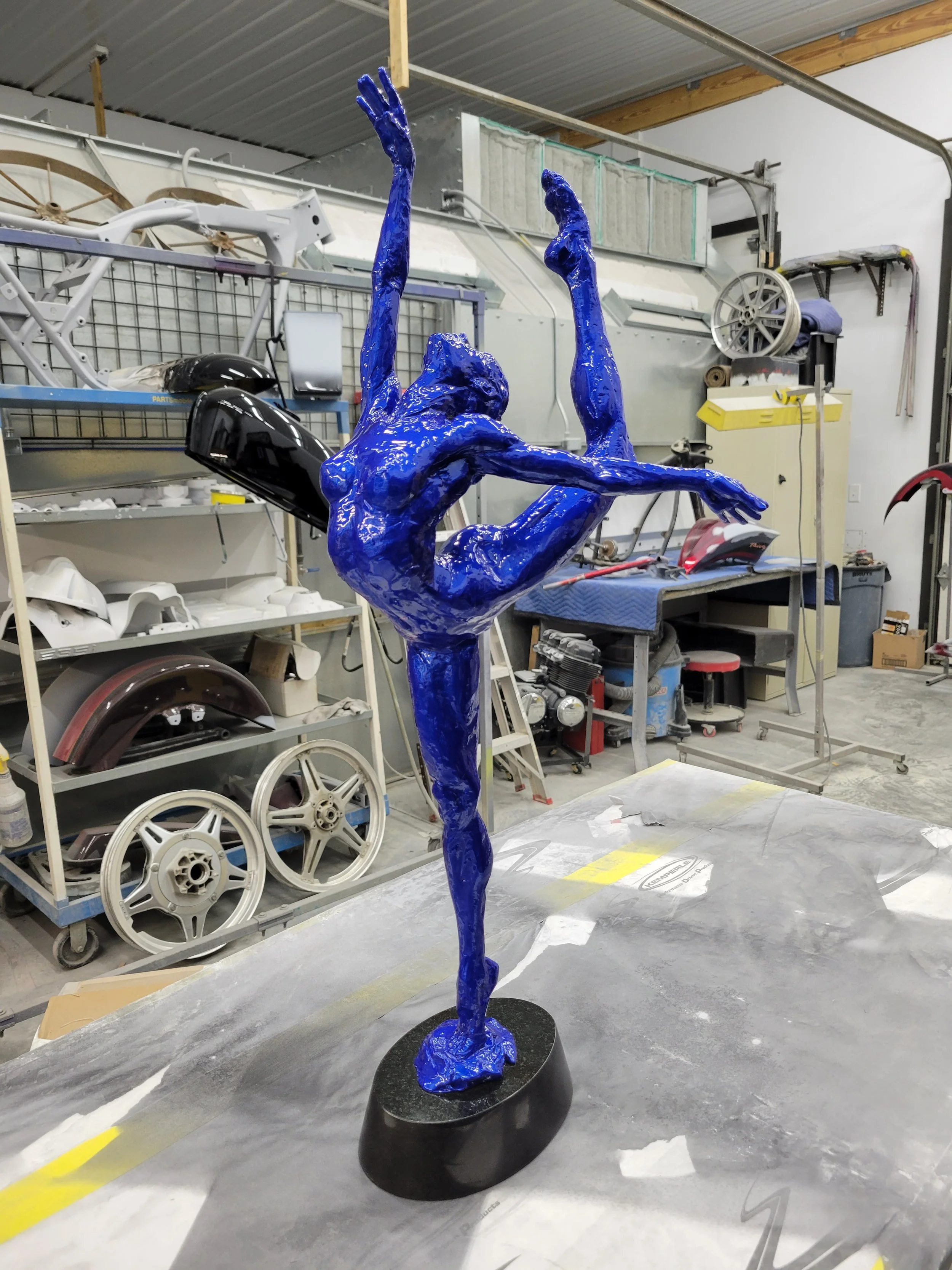 A shiny blue sculpture of a ballet dancer in a dance pose, standing on one leg with arms raised and extended, in a workshop environment.