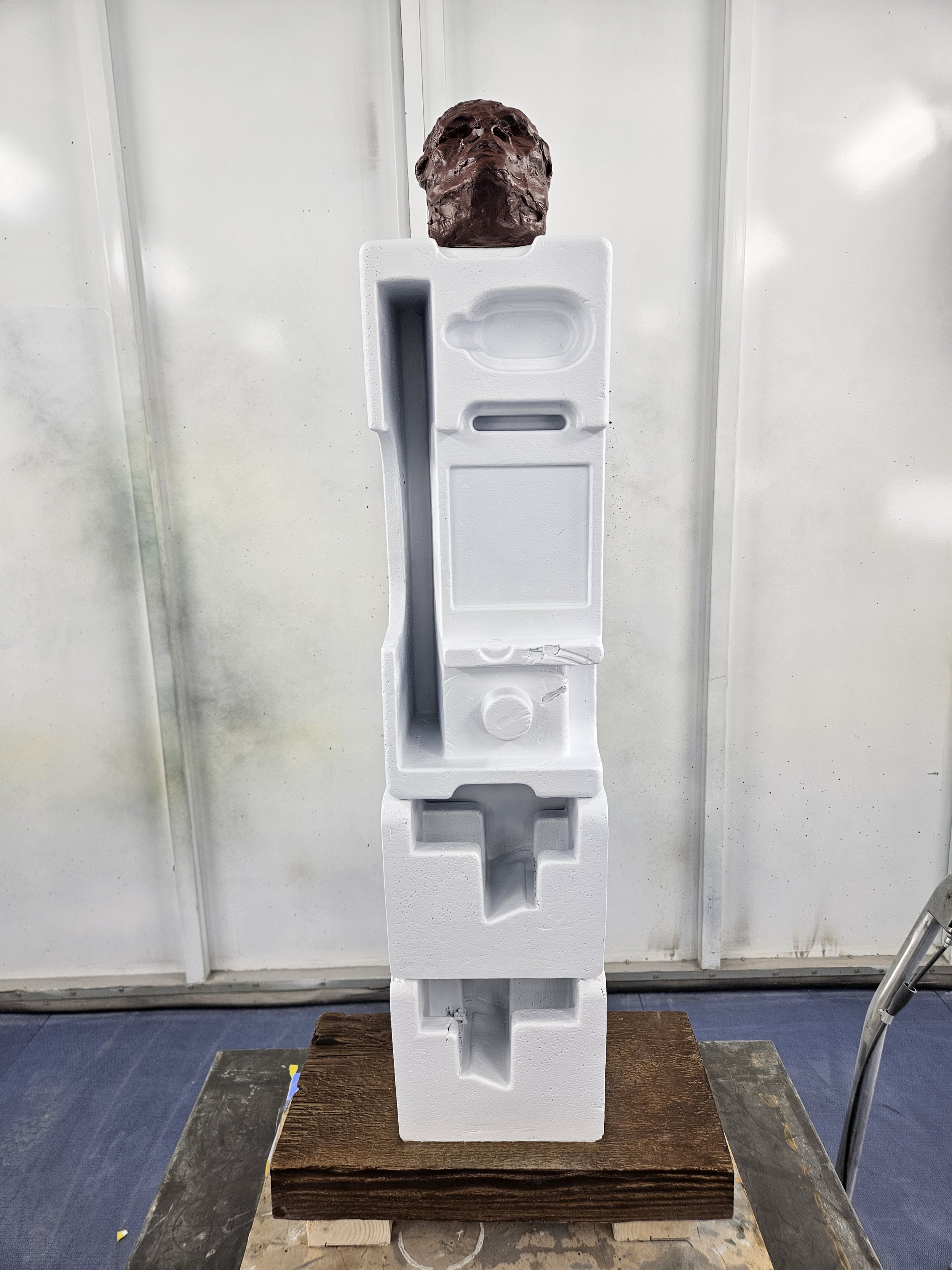Sculpture of a person’s head and shoulders on top of a tall, white foam block with various cut-outs, placed on a wooden platform.