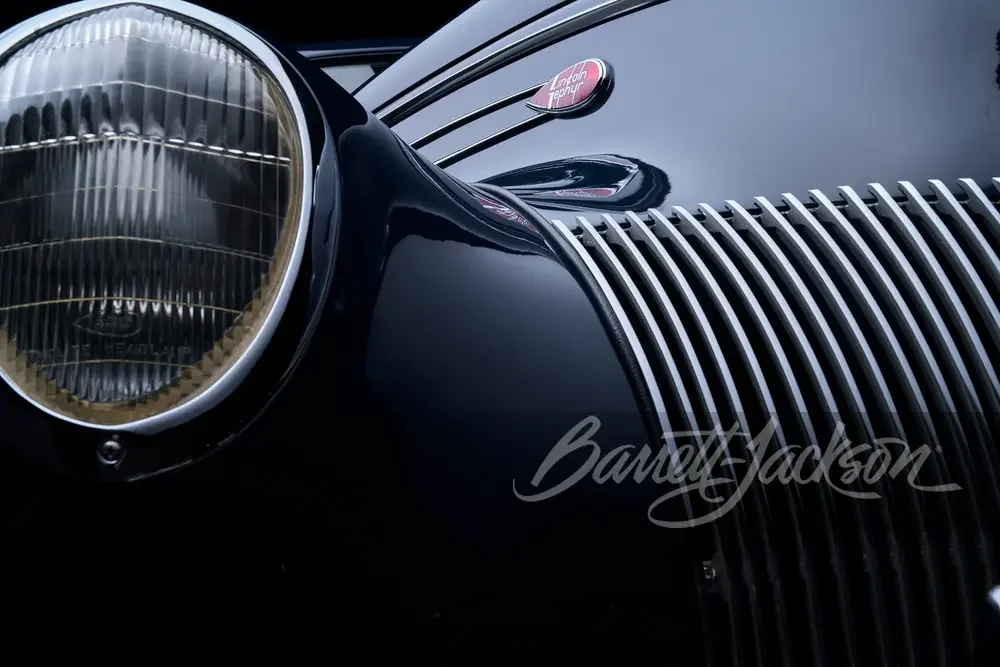 Close-up of the front end of a classic black car showing the large headlight, grille, and a badge on the hood that reads 'Landau Tropy'.