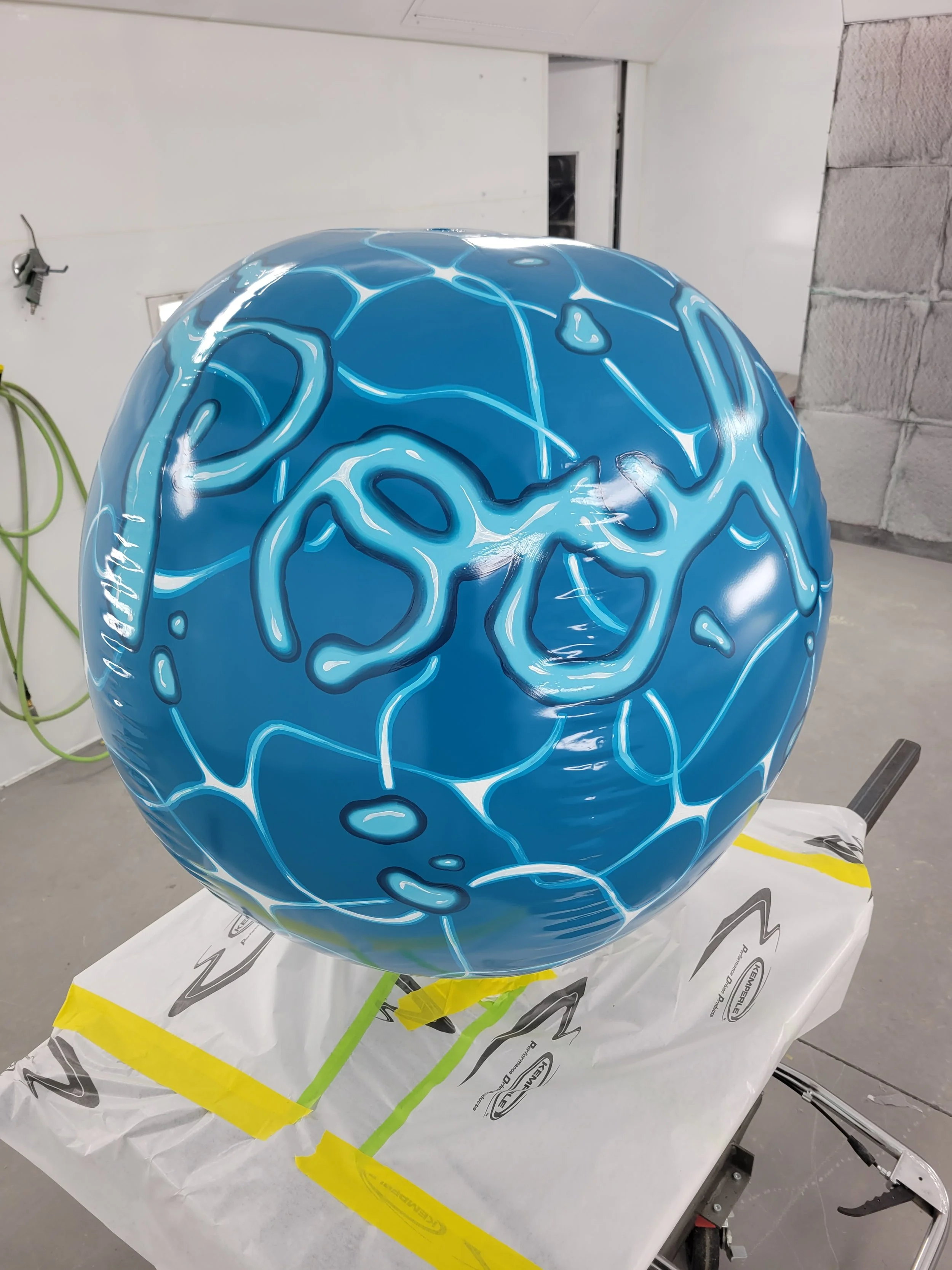 A large blue spherical object with white marbled and crackled patterns, placed on a table covered with white and yellow protective paper in a workshop or garage setting.