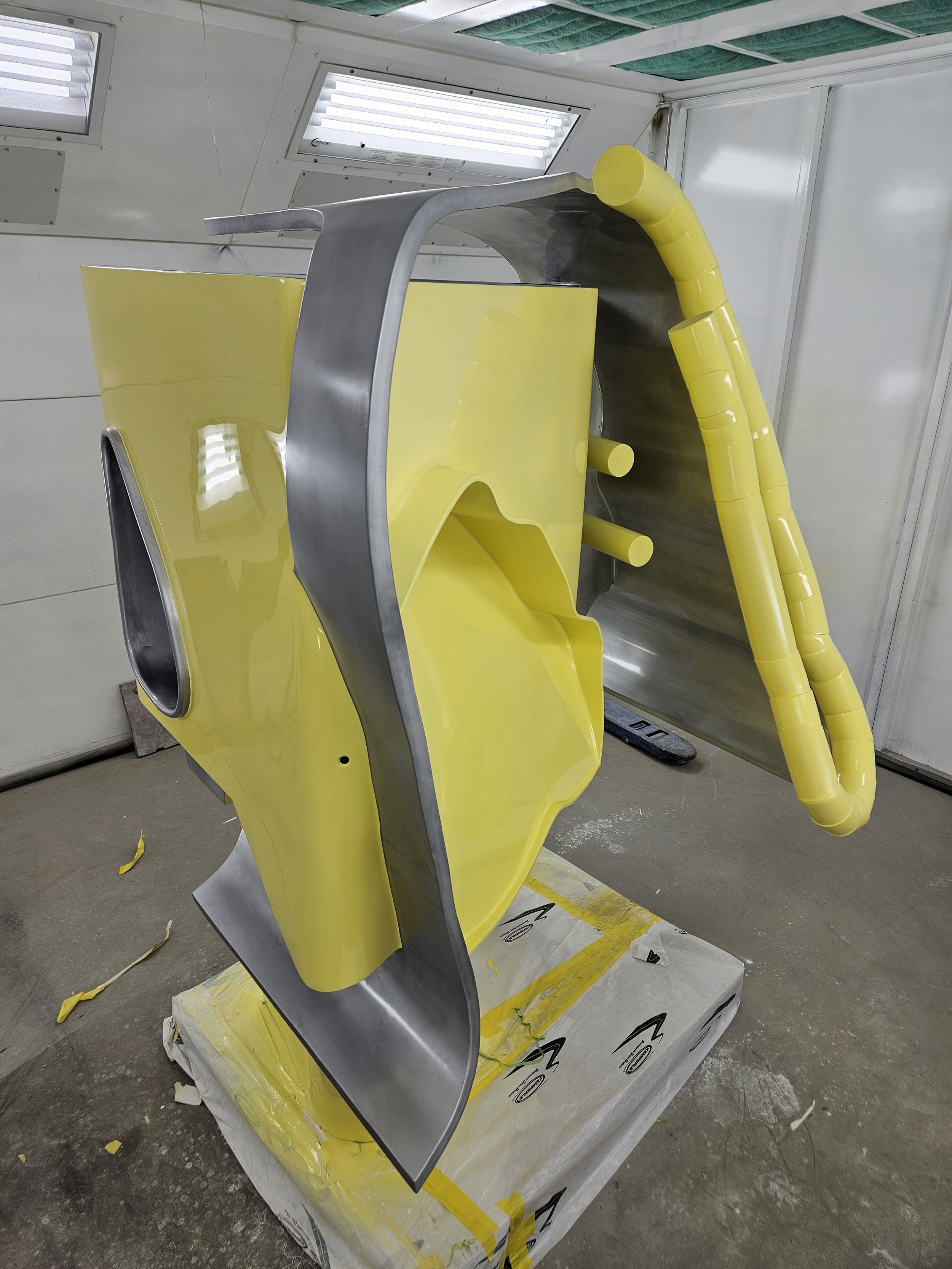 A partially assembled yellow and gray spacecraft or satellite with a curved yellow tube, set on a white platform with yellow tape in a white-walled workshop.