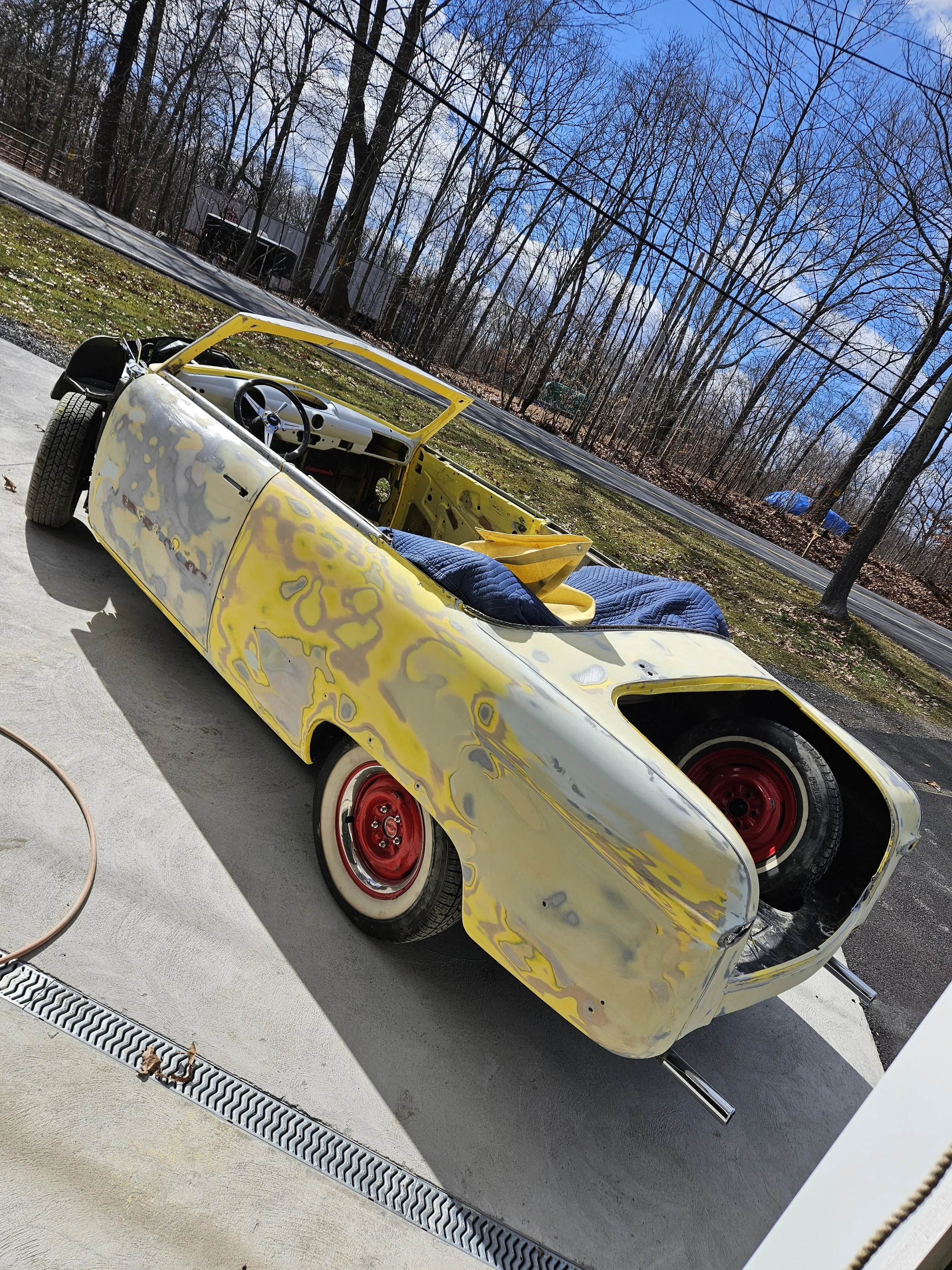 Photo of a vintage convertible car with a yellow primer coat, red wheels, and some patches of exposed metal, parked on a concrete driveway with a blue sky and leafless trees in the background.