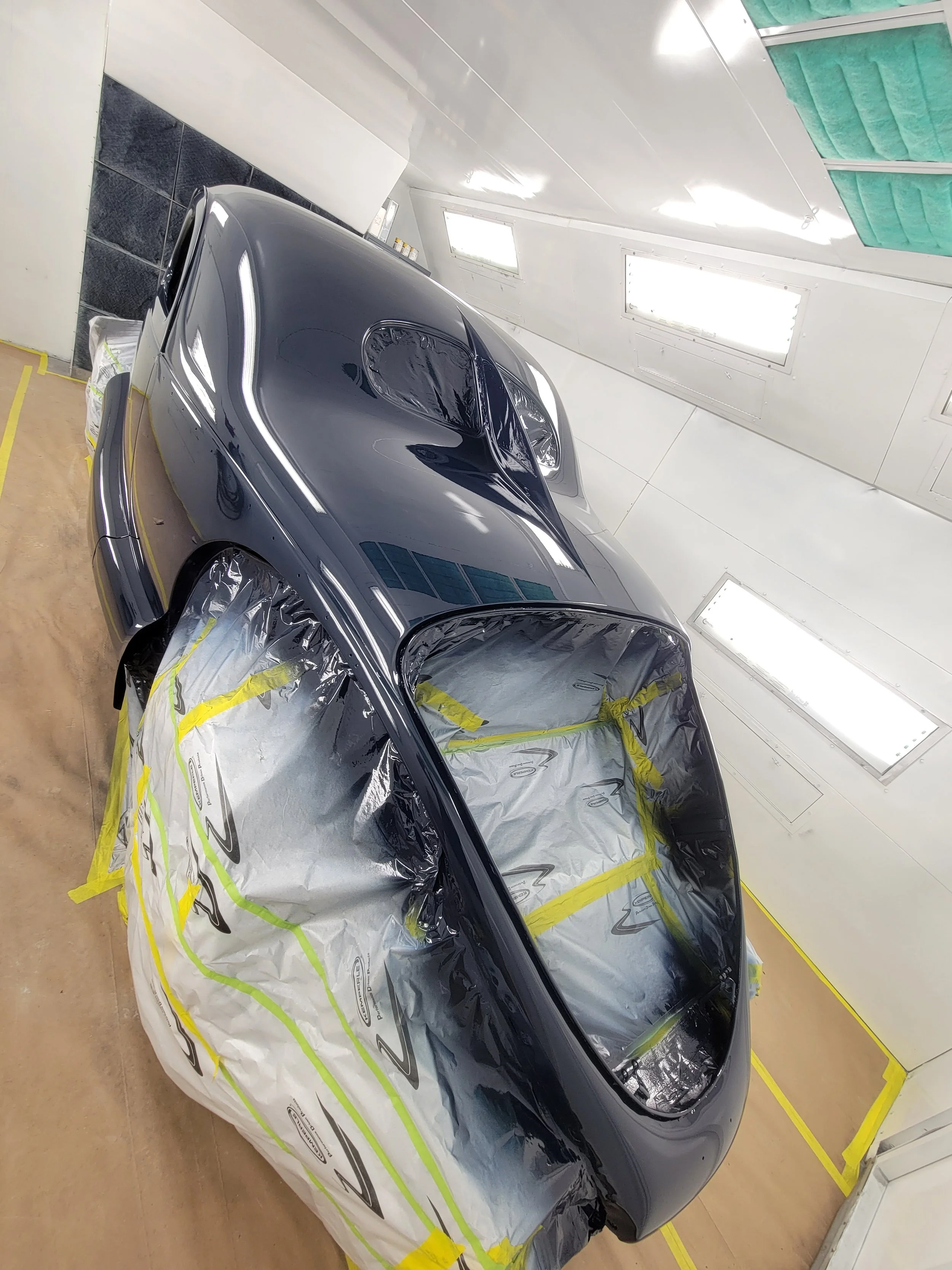 Black sports car being painted inside a spray booth, masked off with paper and plastic covering