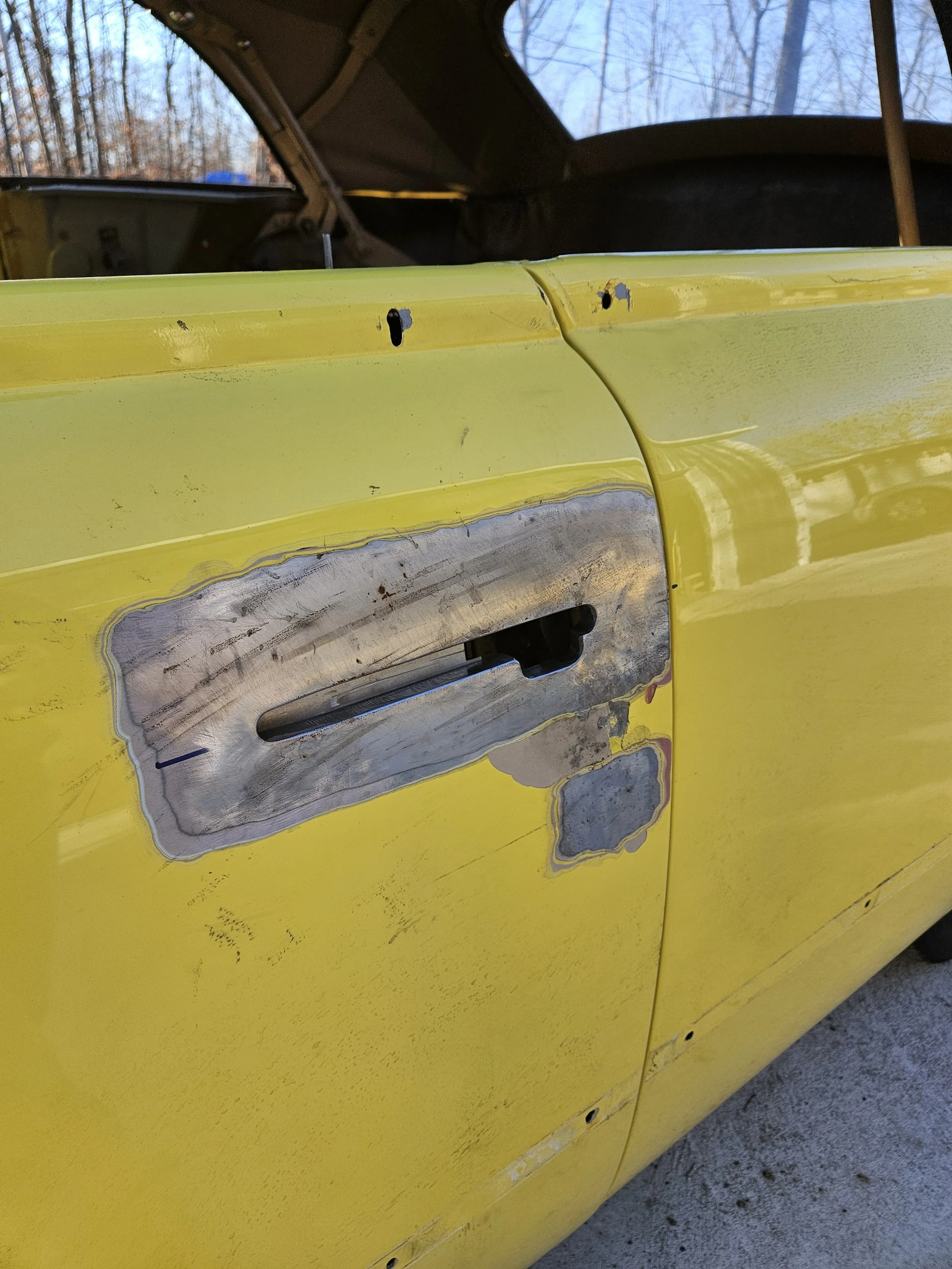 Close-up of a yellow car door with the door handle removed, revealing the metal and primer underneath, and some scratches and dents.