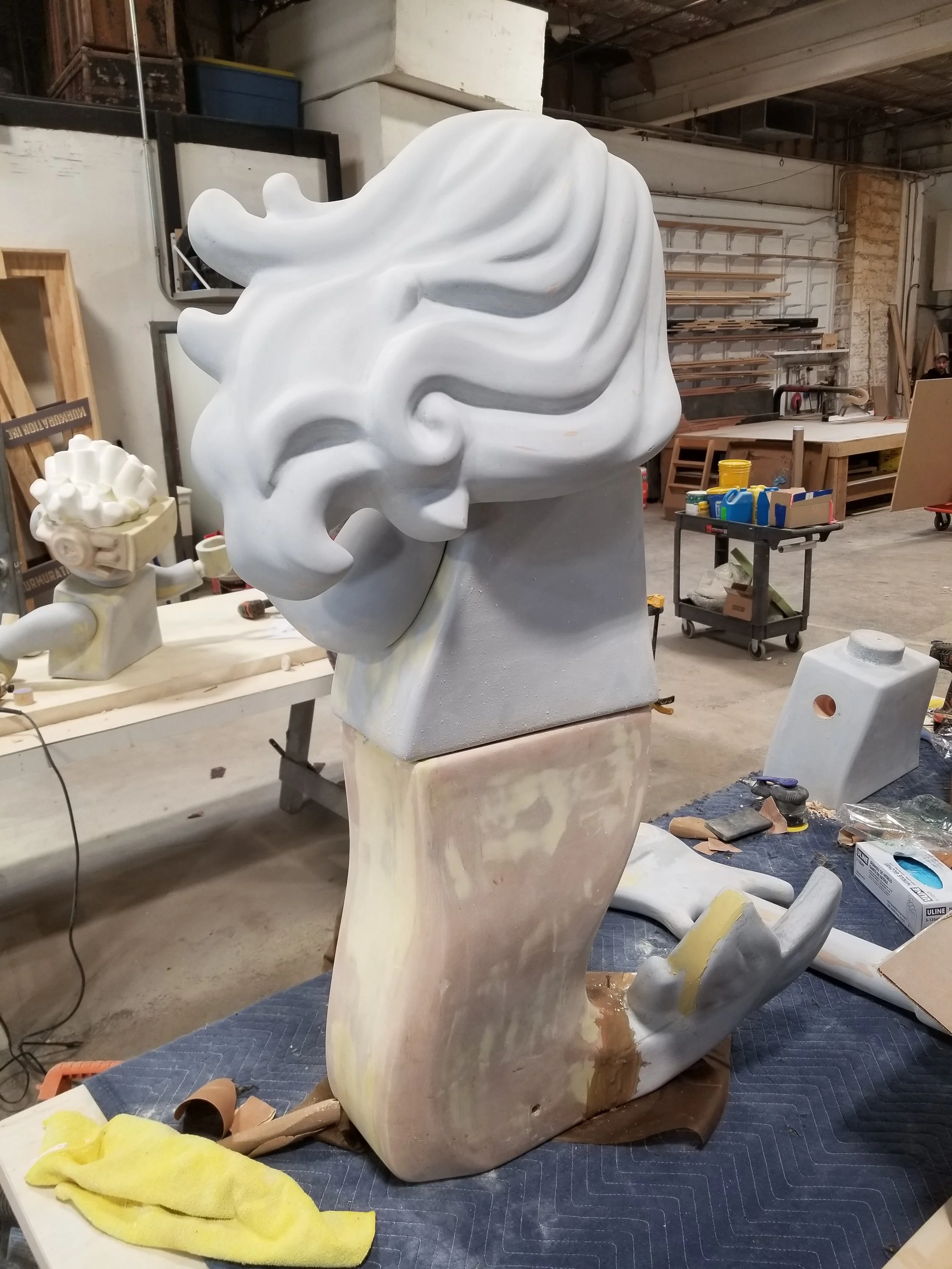 Large LEGO sculpture of a mermaid with flowing hair, mounted on an abstract base in a workshop.