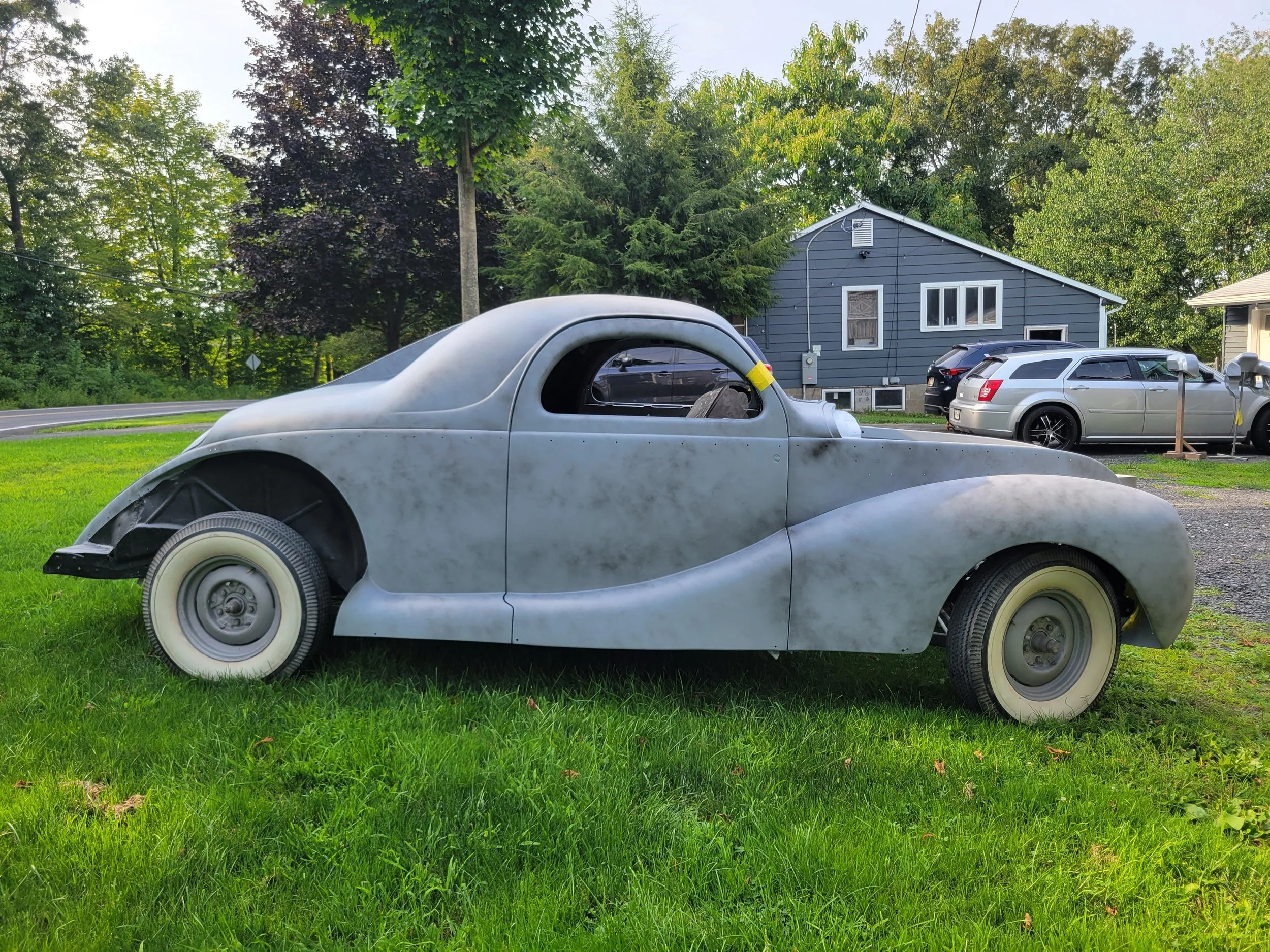 An old, weathered vintage car with missing wheels is parked on a grassy lawn in front of a house, with several modern cars parked nearby.