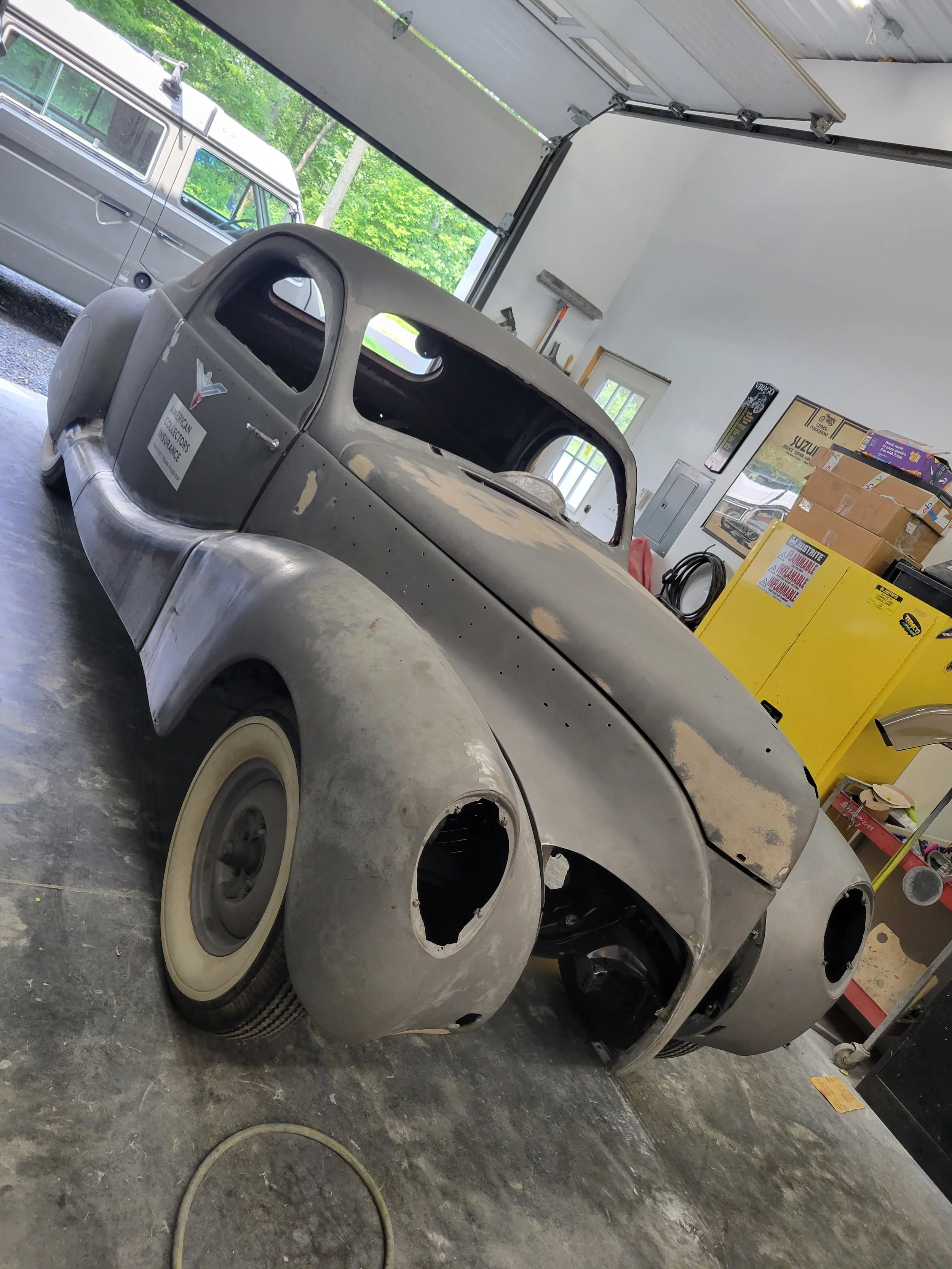 An unfinished vintage car in a garage, with a rough metal body, missing its headlights, and a bubble windshield. The garage contains tools, boxes, and a yellow cabinet.