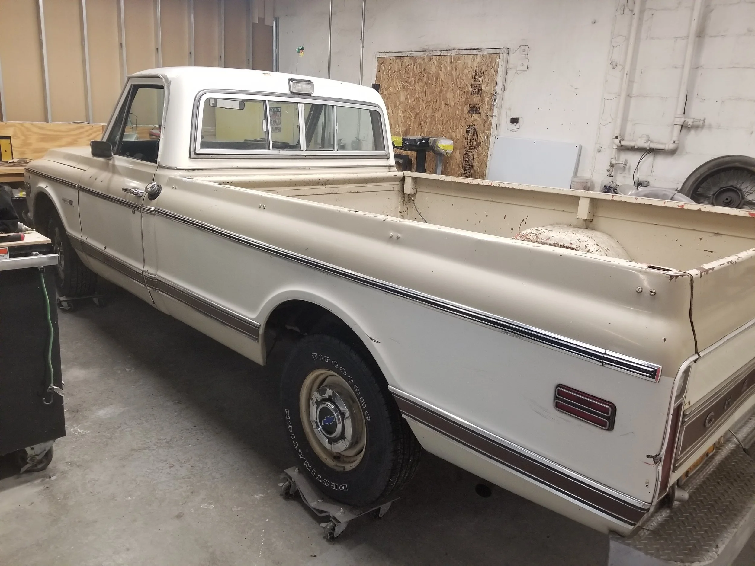 An old cream-colored pickup truck inside a garage, with a wooden door and various tools and equipment around.