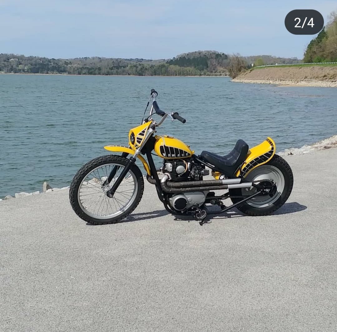 Yellow custom motorcycle parked by a body of water with hills and trees in the background.
