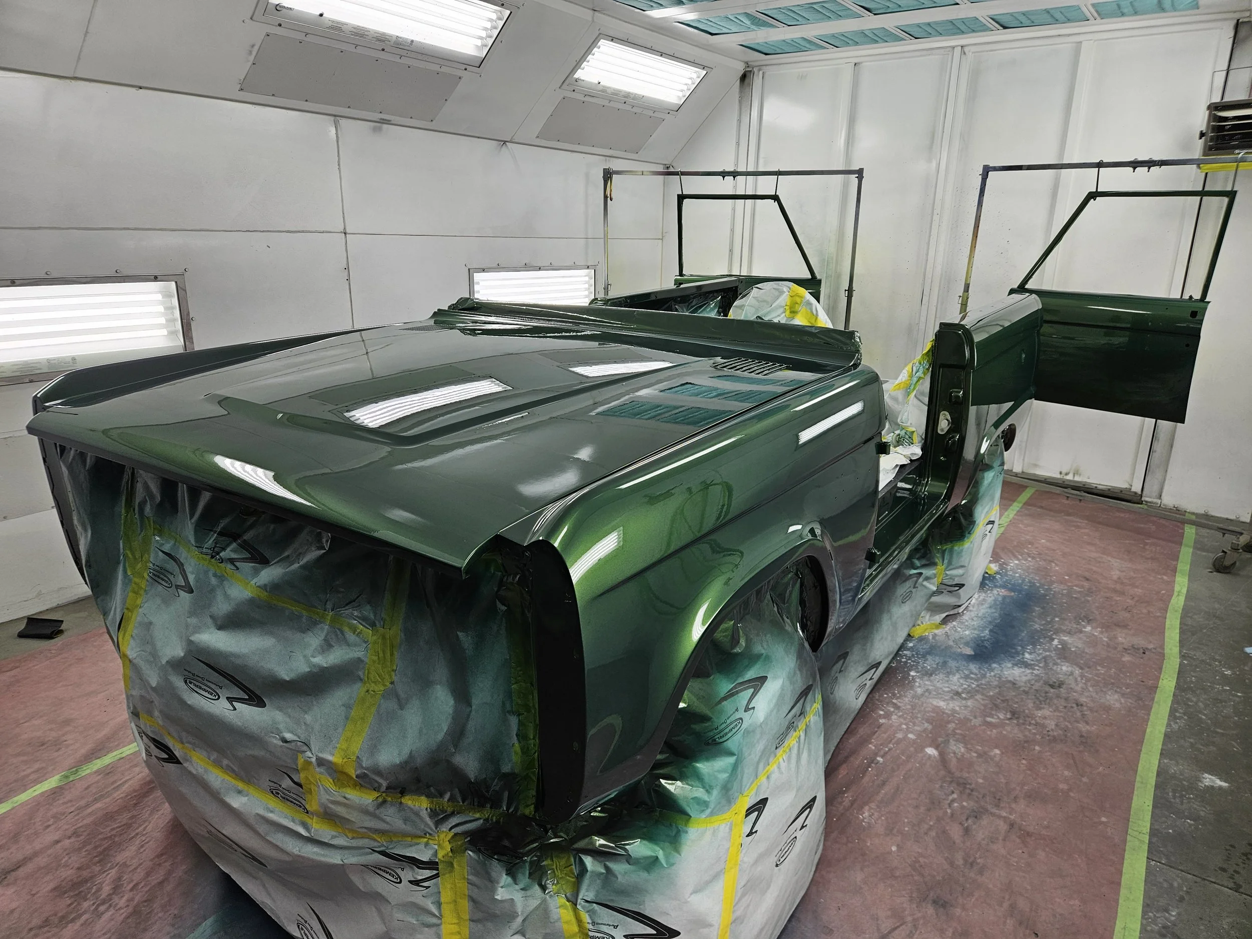 A green sports truck is in a paint booth, partially masked with spray paint on the body.