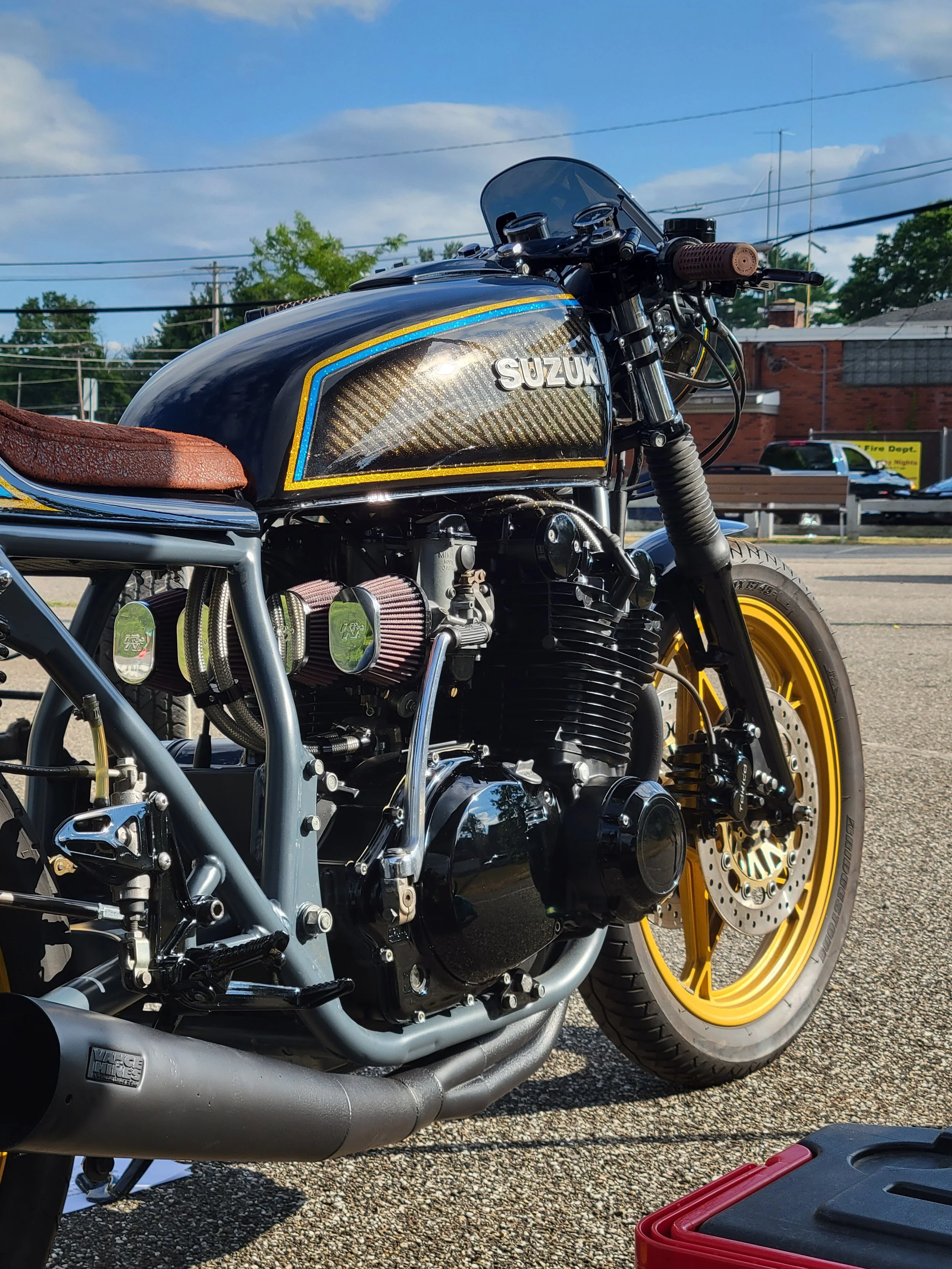Close-up of a custom black Suzuki motorcycle with yellow rims parked outdoors on a sunny day.