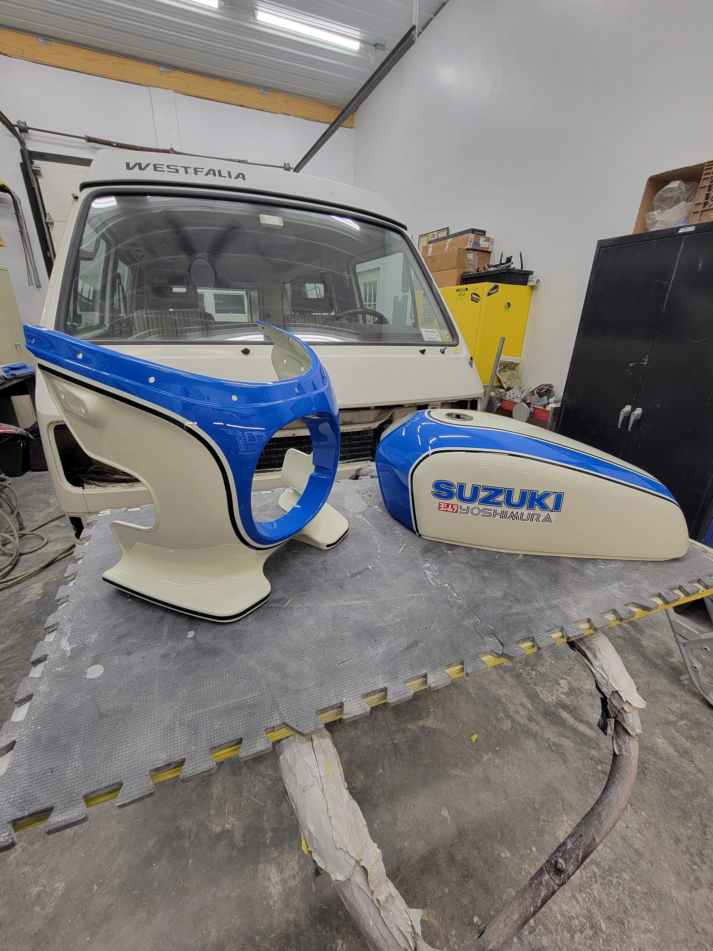 Disassembled motorcycle tank and front cowl painted in blue and white, with 'Suzuki' and 'Yoshimura' logos, placed on workbench in workshop.