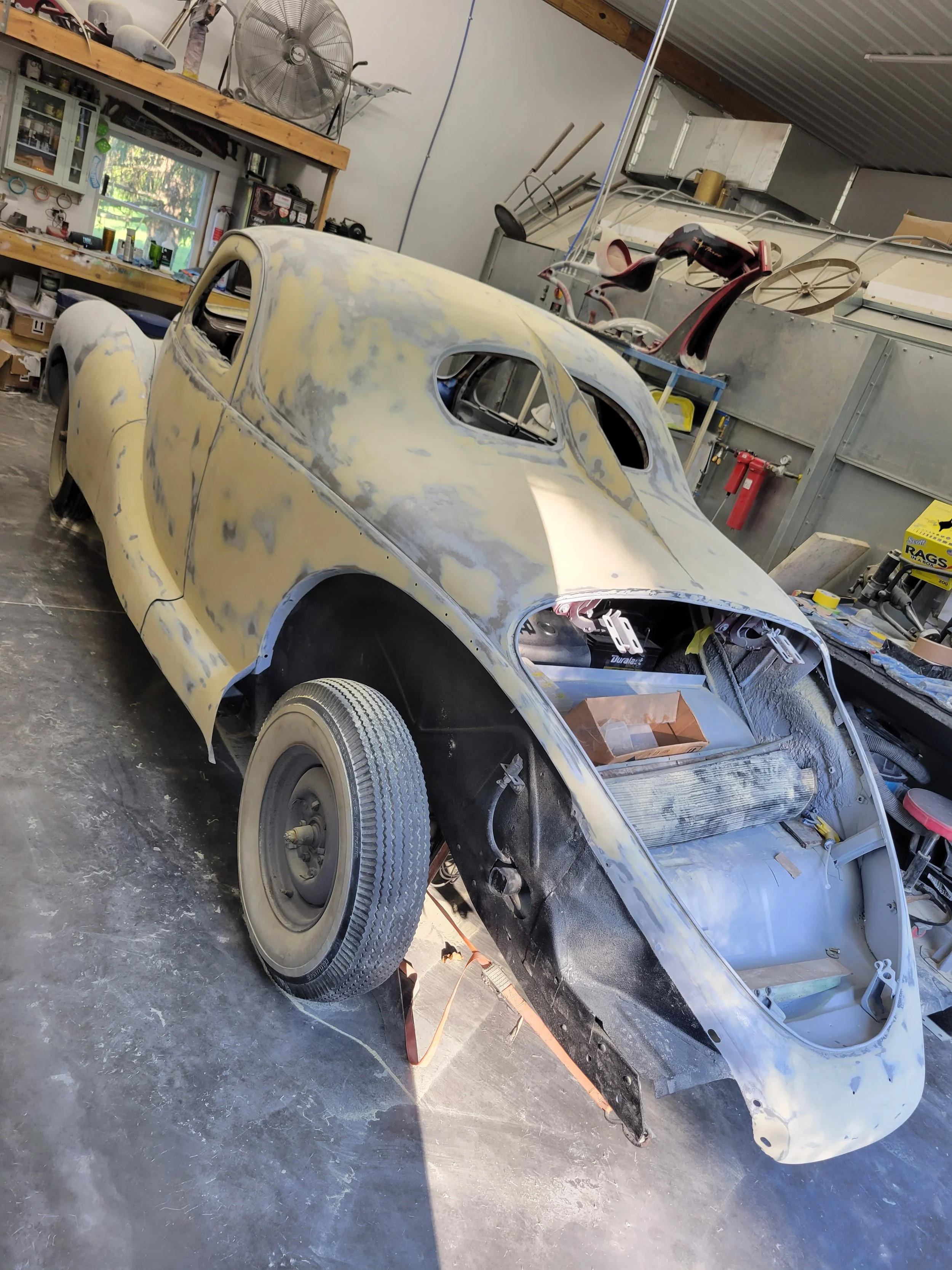 Unfinished vintage car body in a garage workspace, with unpainted surface, missing engine, and surrounded by tools and parts.