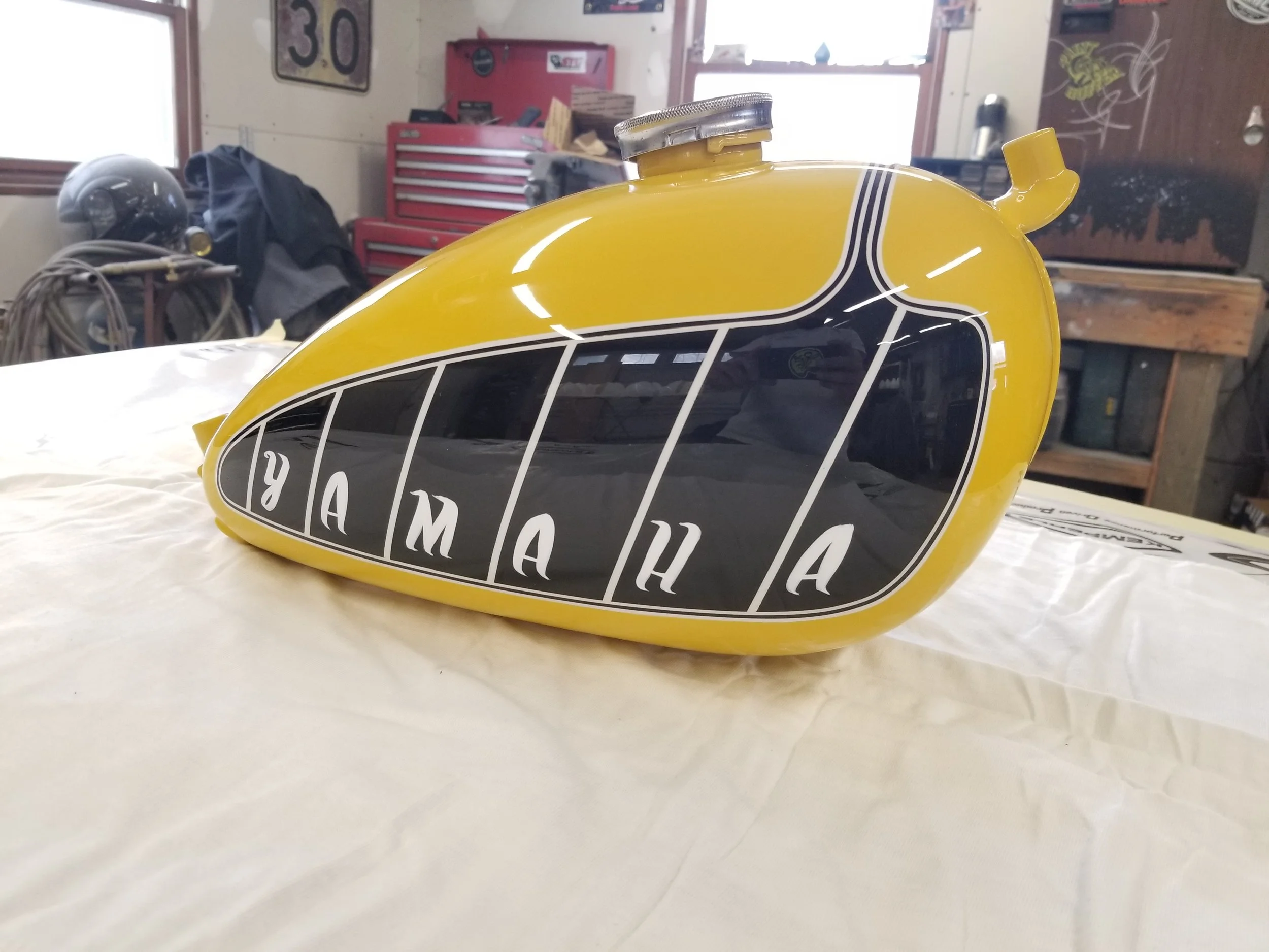 Yellow Yamaha gas tank with Yamaha logo in black and white, placed on a table in a workshop with tools and equipment in the background.