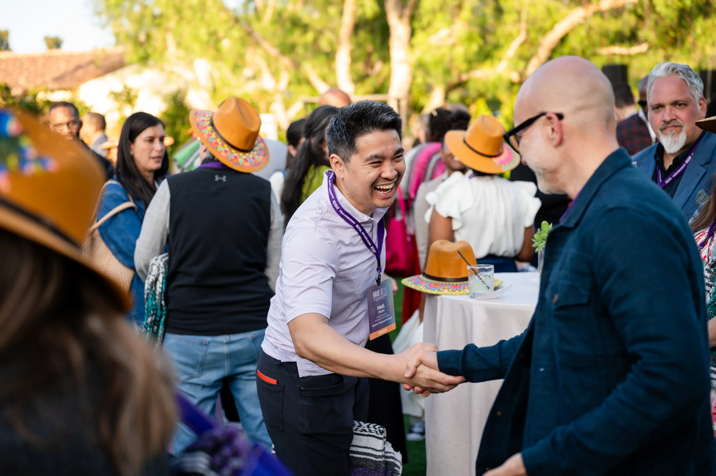 Two men shaking hands at an outdoor event with a crowd of people in the background. One man is smiling widely, wearing a light-colored shirt and a lanyard. The other man is bald, wearing glasses, and a dark jacket. Several people are wearing sombrero