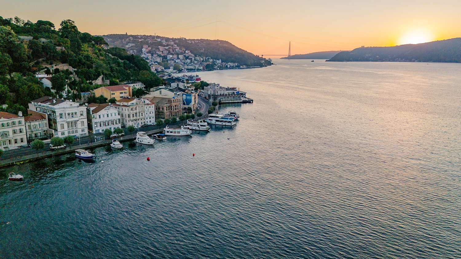 A waterfront view at sunset with boats docked along the harbor, residential buildings in the foreground, a hillside covered with trees, and a bridge in the distance over the water.