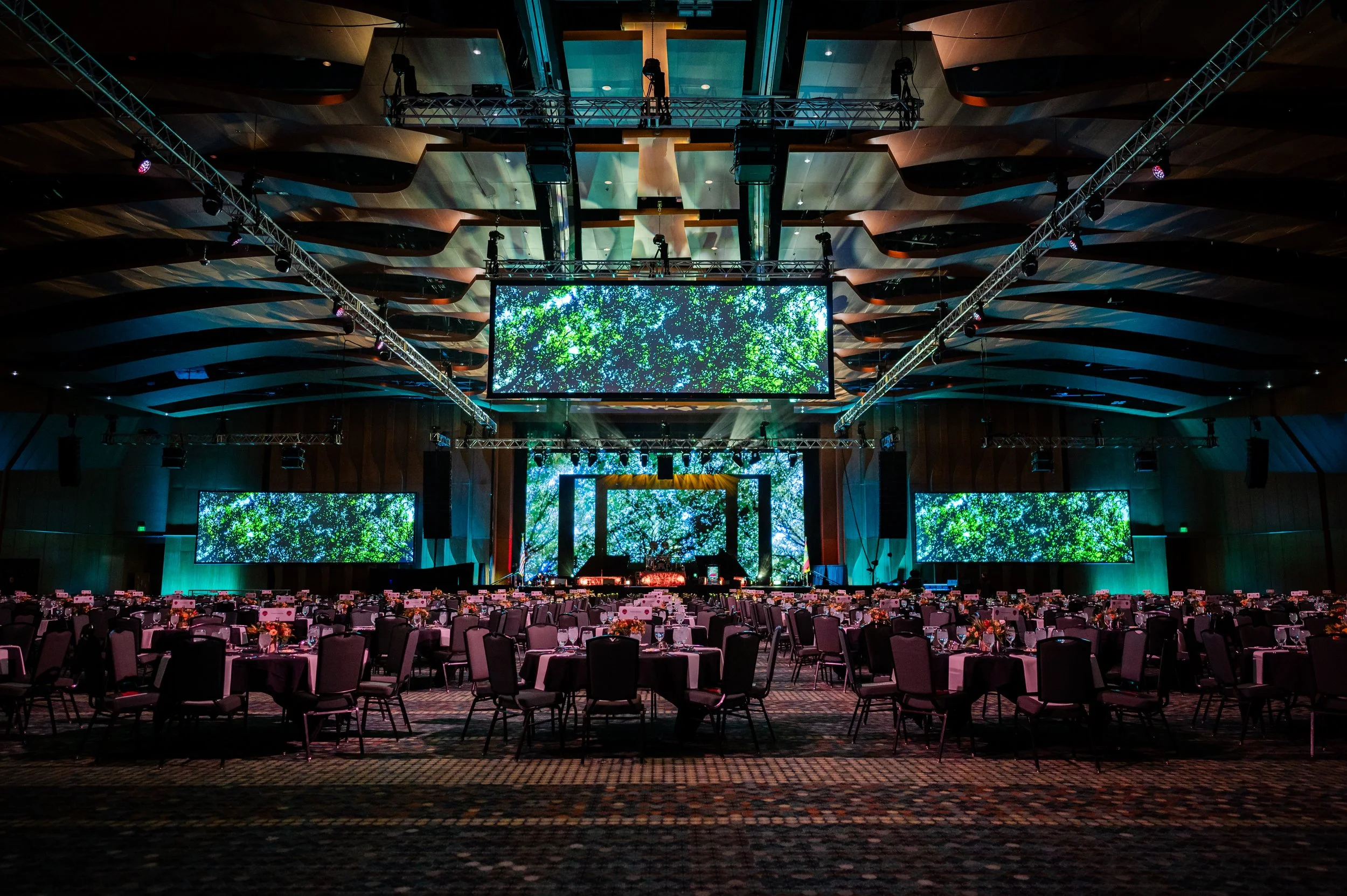 Large banquet hall decorated for an event with waterfalls or nature-themed digital screens, round tables with chairs, and stage setup with lighting and audio equipment.