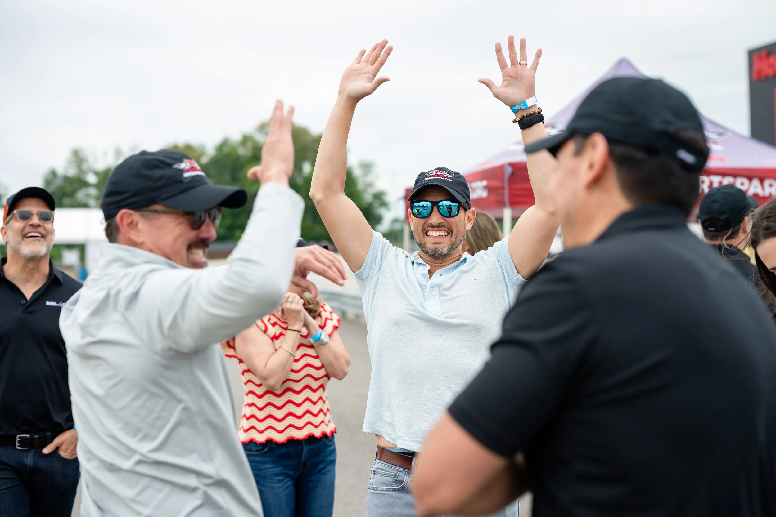 Group of people outdoors at a celebration, with a man in a gray polo and sunglasses raising his hands in the air while people around him smile and cheer.