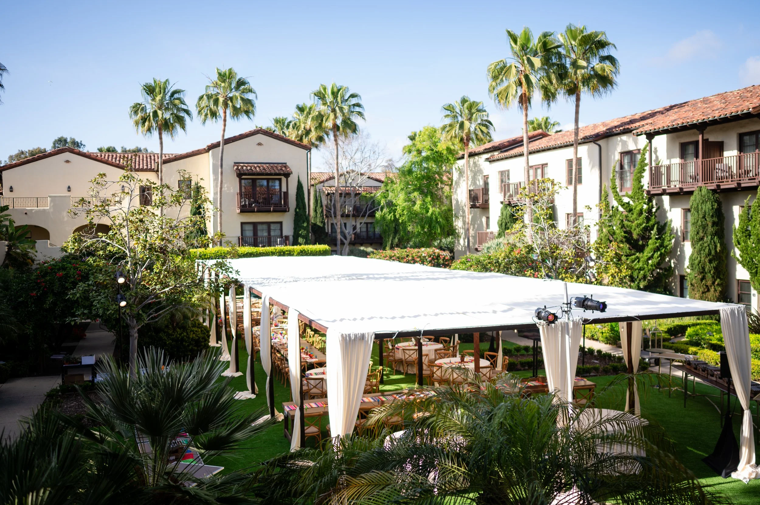 Outdoor event setup with a large white canopy, wooden tables, and chairs surrounded by lush greenery and tall palm trees, in a residential complex with white buildings and red-tiled roofs.