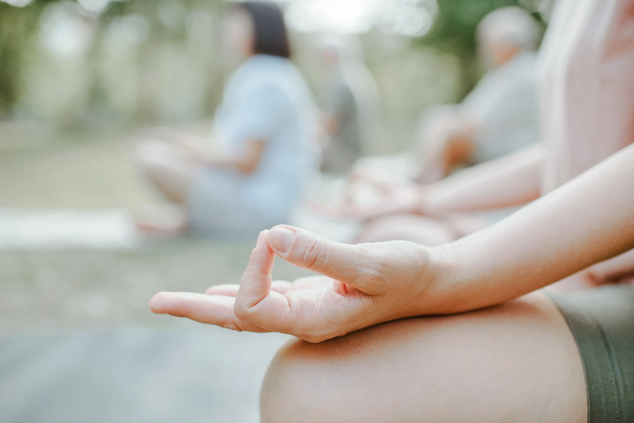 Close-up of a person's hand with fingers in a yoga mudra, resting on their leg outdoors, with blurred background of other people and trees.
