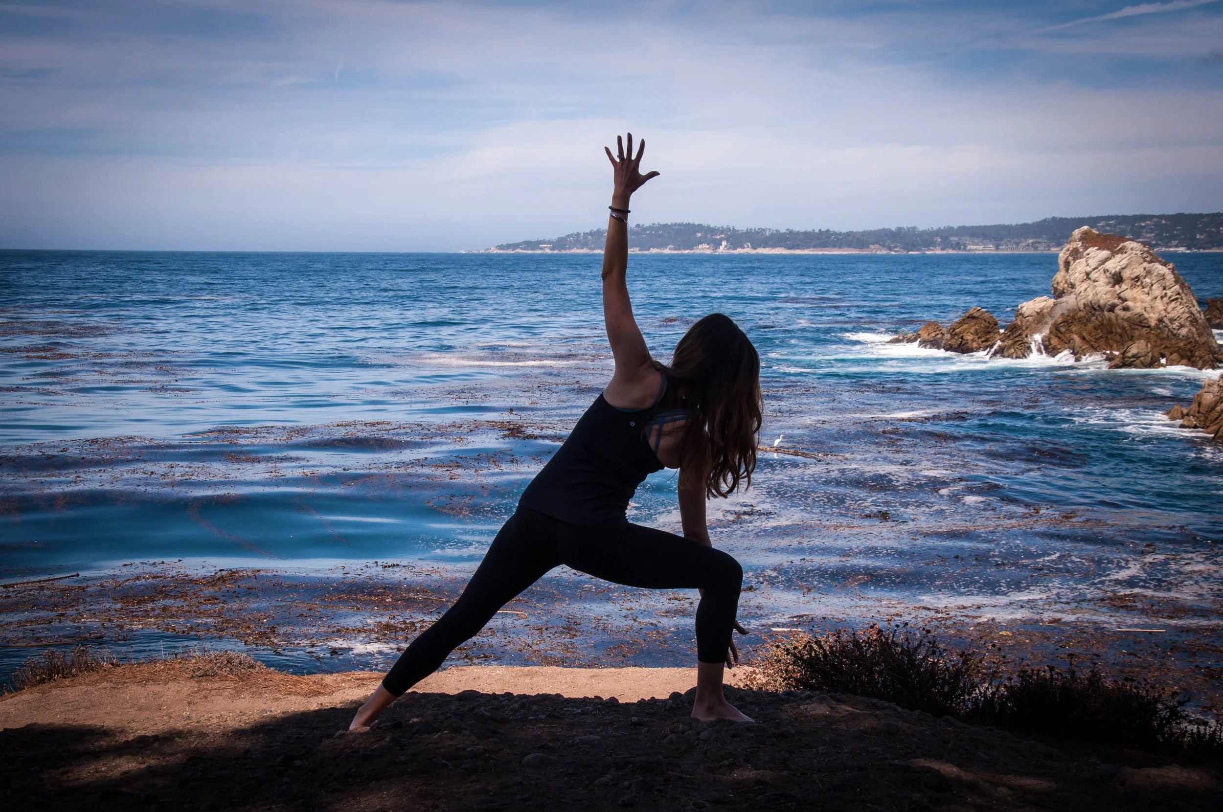 A woman practicing yoga on a beach, performing a triangle pose with one arm extended upward and the other reaching toward the ground, with rocks and the ocean in the background.