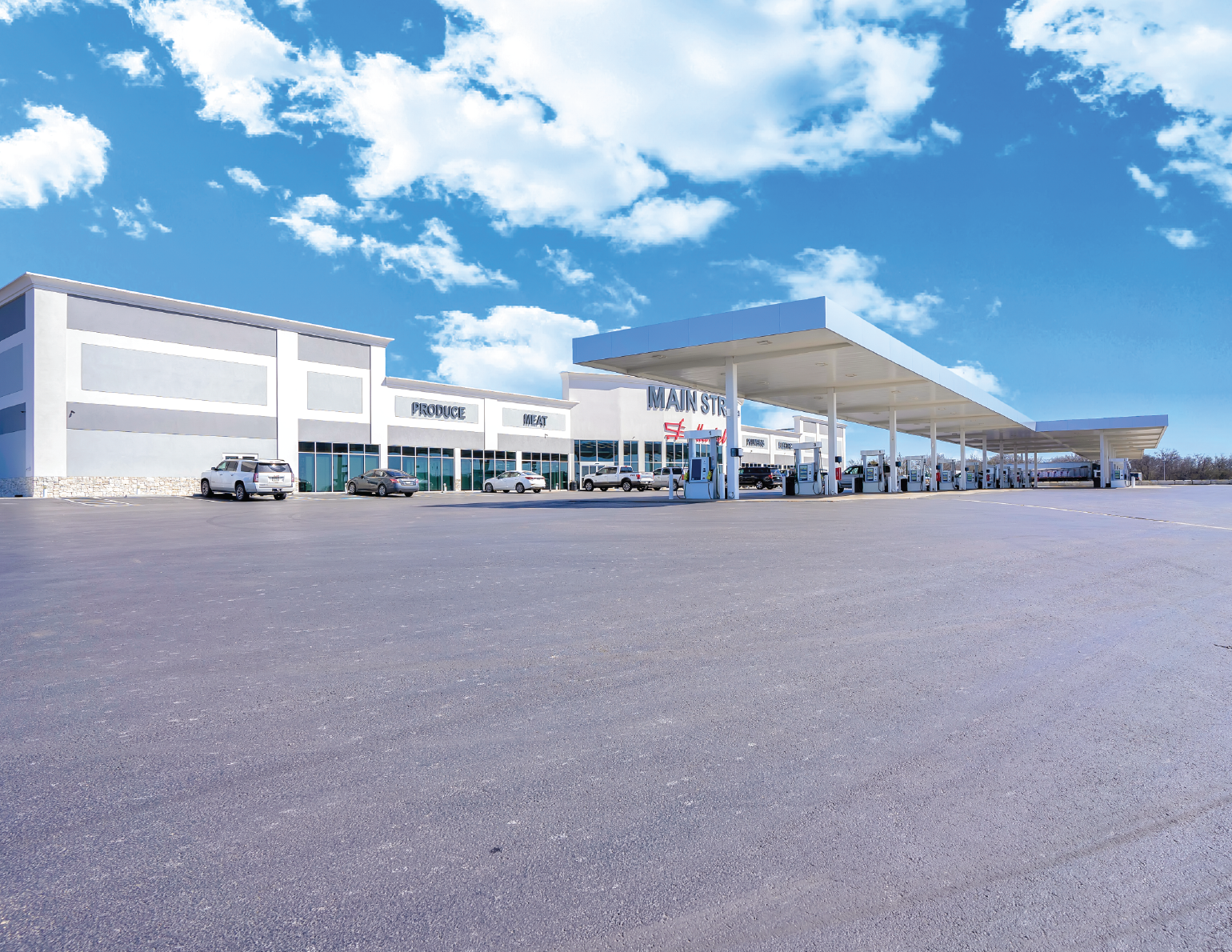 Empty parking lot outside a modern gas station with a convenience store. Several cars are parked, and gas pumps are visible under a large canopy. The store has signs for produce, meat, and main street.