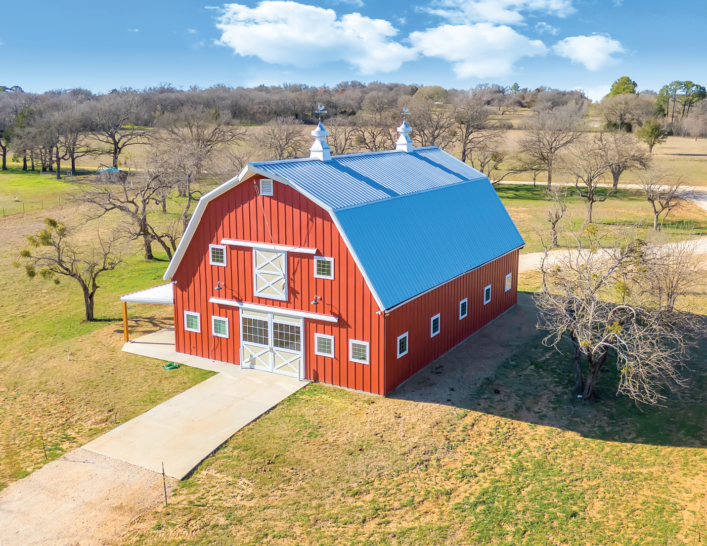 A large red barn with white trim and a blue metal roof situated in a rural landscape with leafless trees, grassy fields, and a partly cloudy sky.