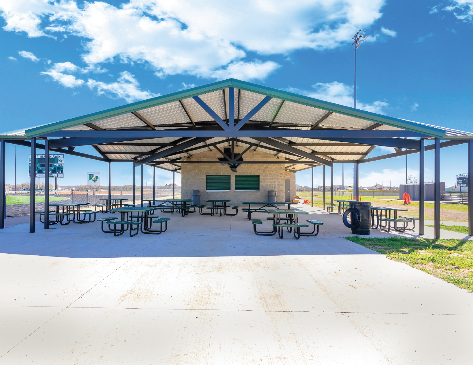 Empty outdoor picnic shelter with multiple tables and benches, trash cans, and a small structure with green shutters under a blue sky with scattered clouds.