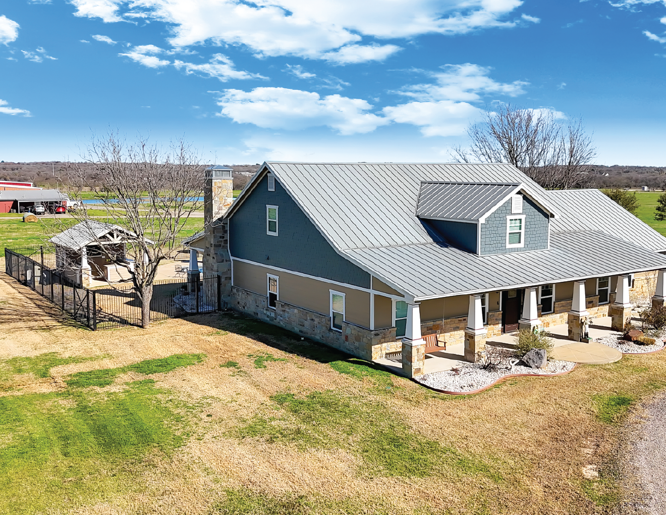 A large, modern house with a metal roof, stone and siding exterior, porch with columns, and a backyard with a tree and a fenced area, set against a bright blue sky with some clouds.