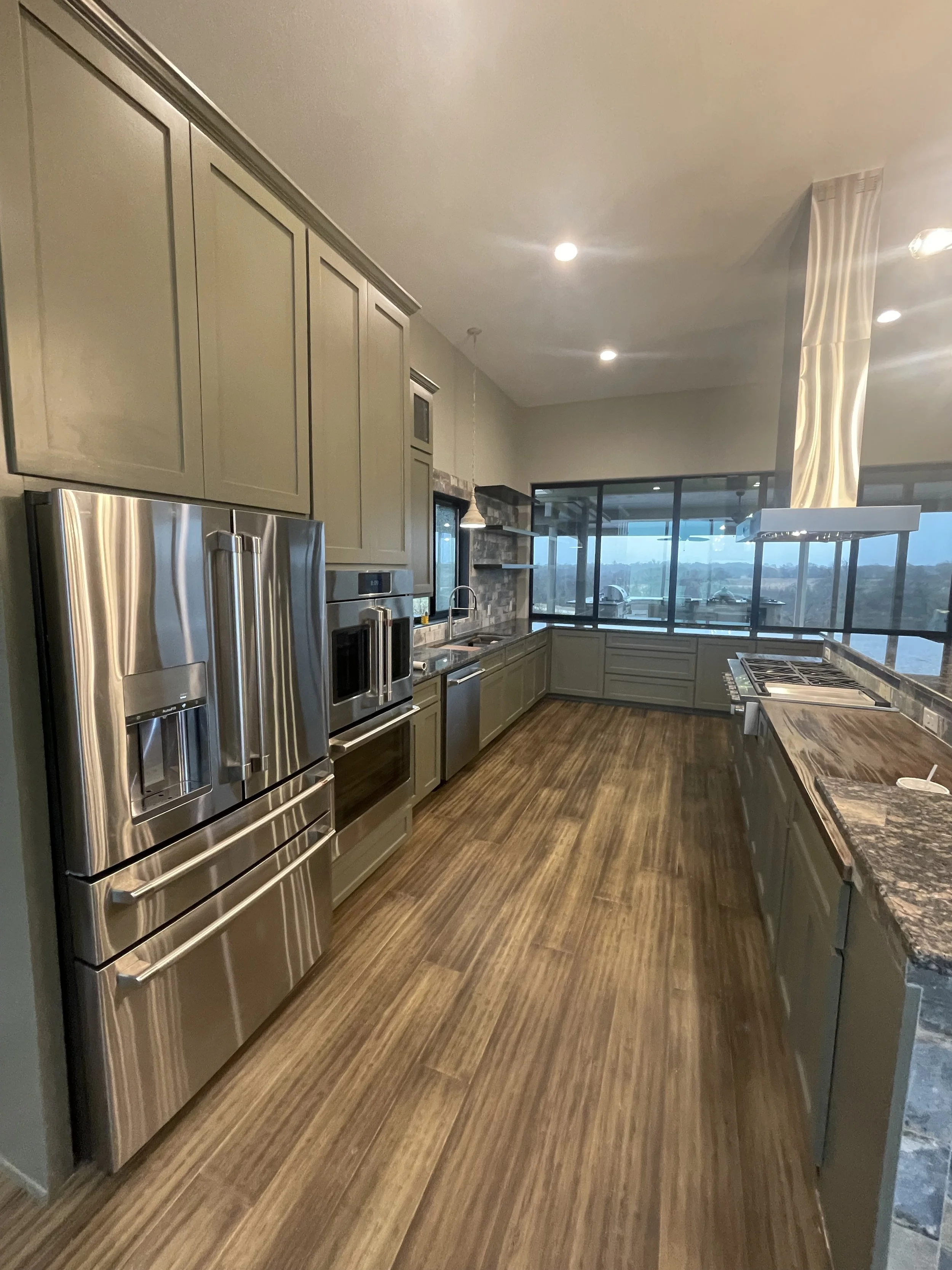 Modern kitchen with gray cabinets, stainless steel refrigerator, and wooden flooring, featuring large windows and granite countertops.