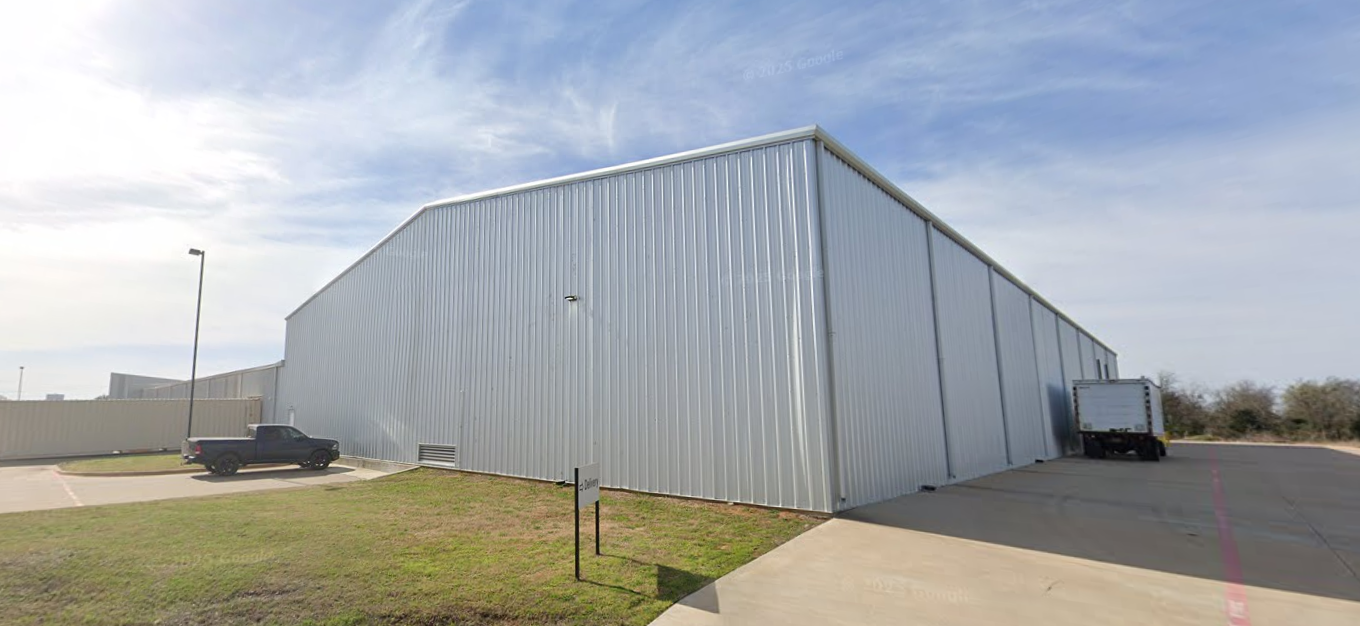Large metal industrial building with a sloped roof, gray exterior, parked black pickup truck, and a delivery truck on a paved lot under partly cloudy sky.