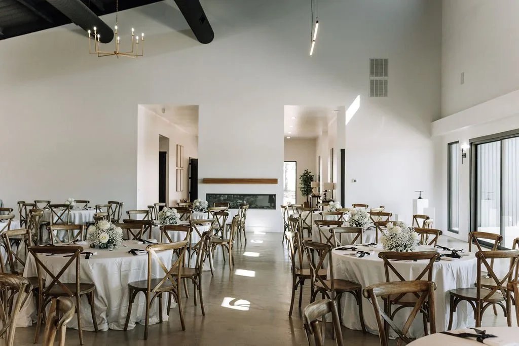 Empty banquet hall decorated with white tablecloths, floral centerpieces, and wooden chairs, with large windows and modern lighting fixtures.