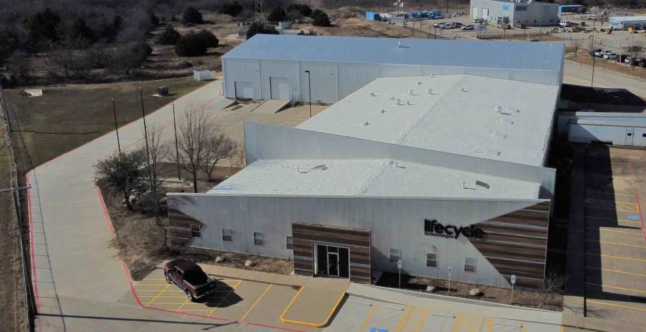 An industrial building labeled Lifecycle, with a white and brown exterior, surrounded by parking lots and some trees, with a clear sky overhead.
