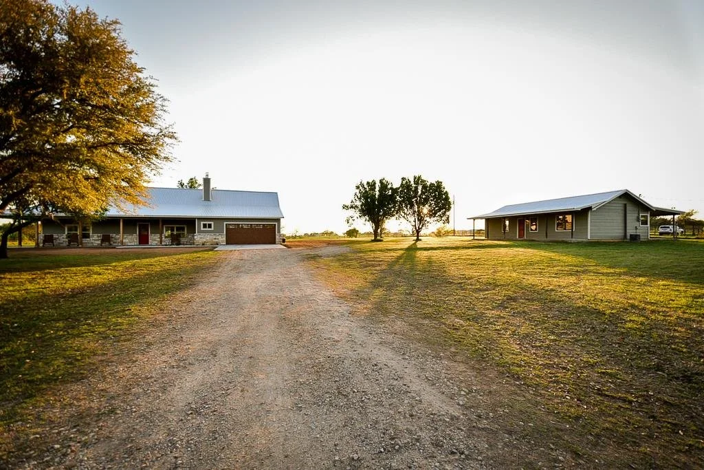 Sunset or sunrise over two houses on a rural property with a dirt driveway and trees.