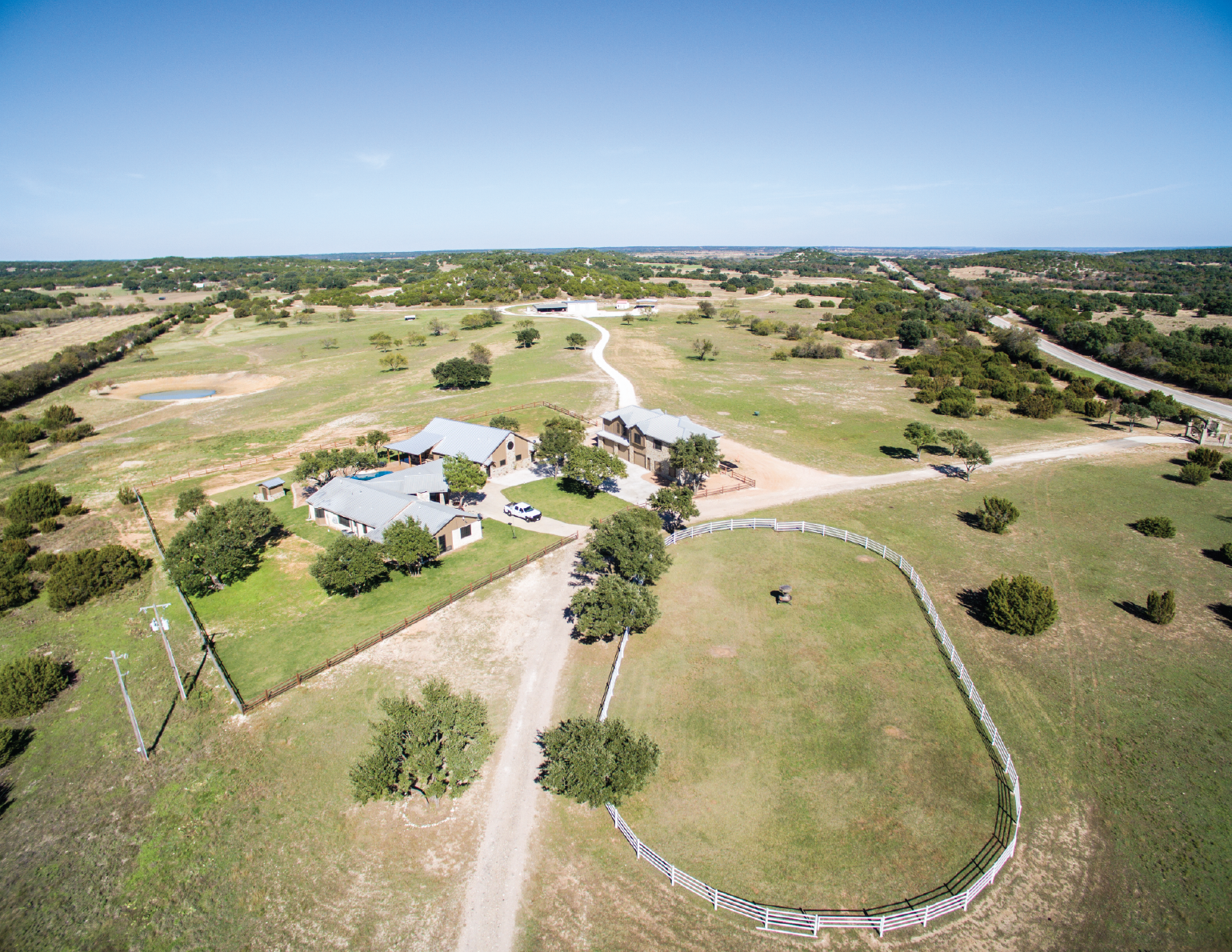 Aerial view of a rural property with multiple buildings, fenced pasture, trees, a pond, gravel paths, and open fields.