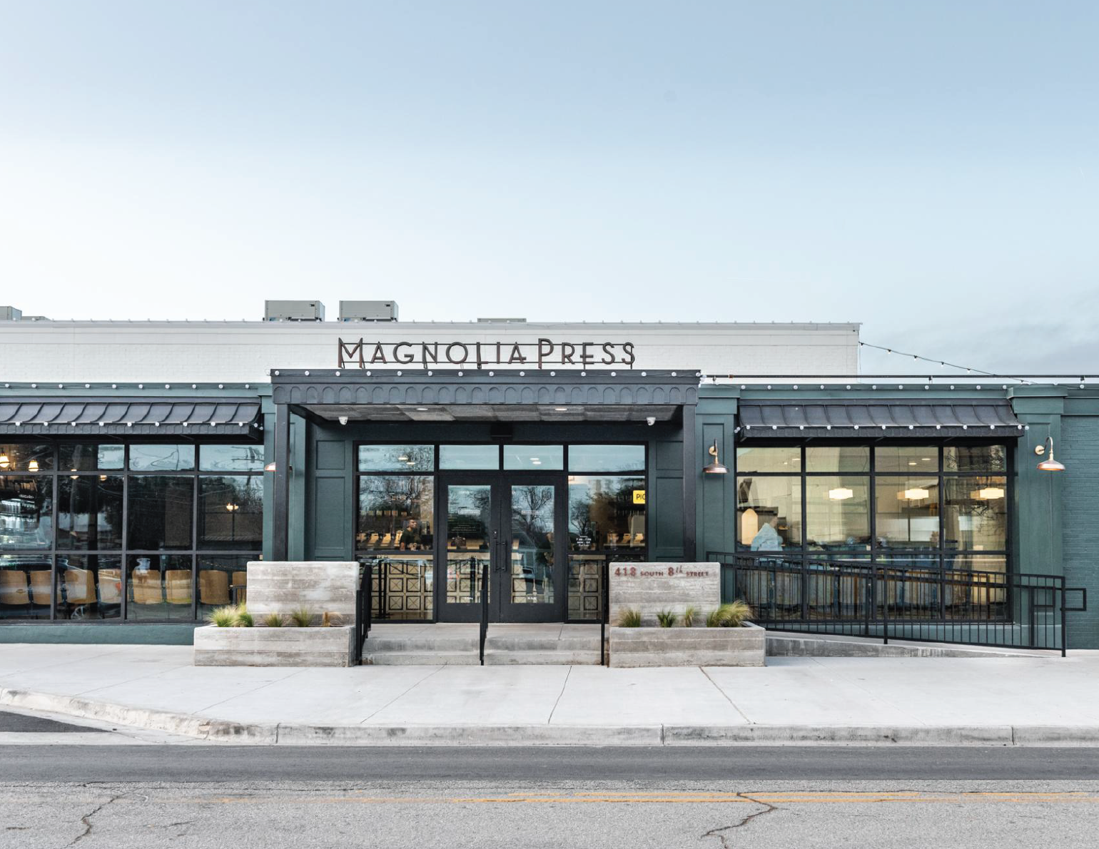 Exterior view of Magnolía Press café with large glass windows, black door, and signage on a cloudy day.
