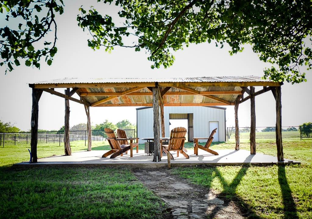 An outdoor pavilion with a metal roof, wooden chairs, and a small shed in a rural setting with green grass and trees.