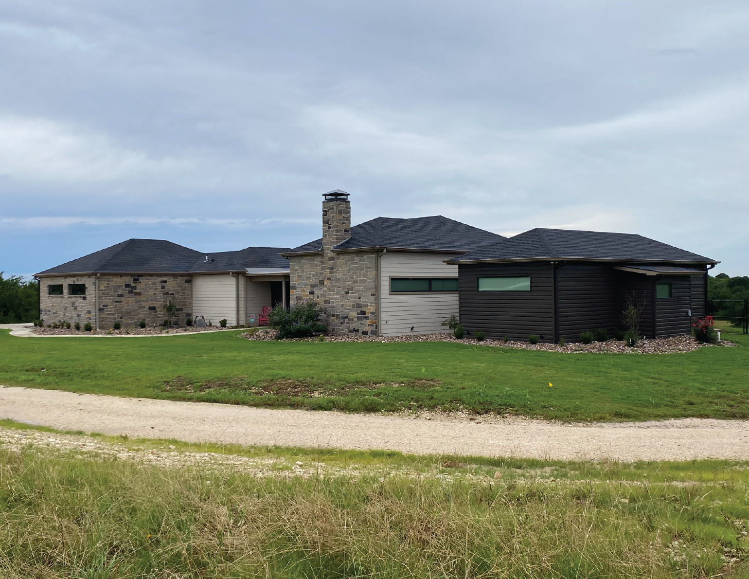 A modern house with a mix of brick and black siding exterior, surrounded by a green lawn and a gravel driveway, under a cloudy sky.