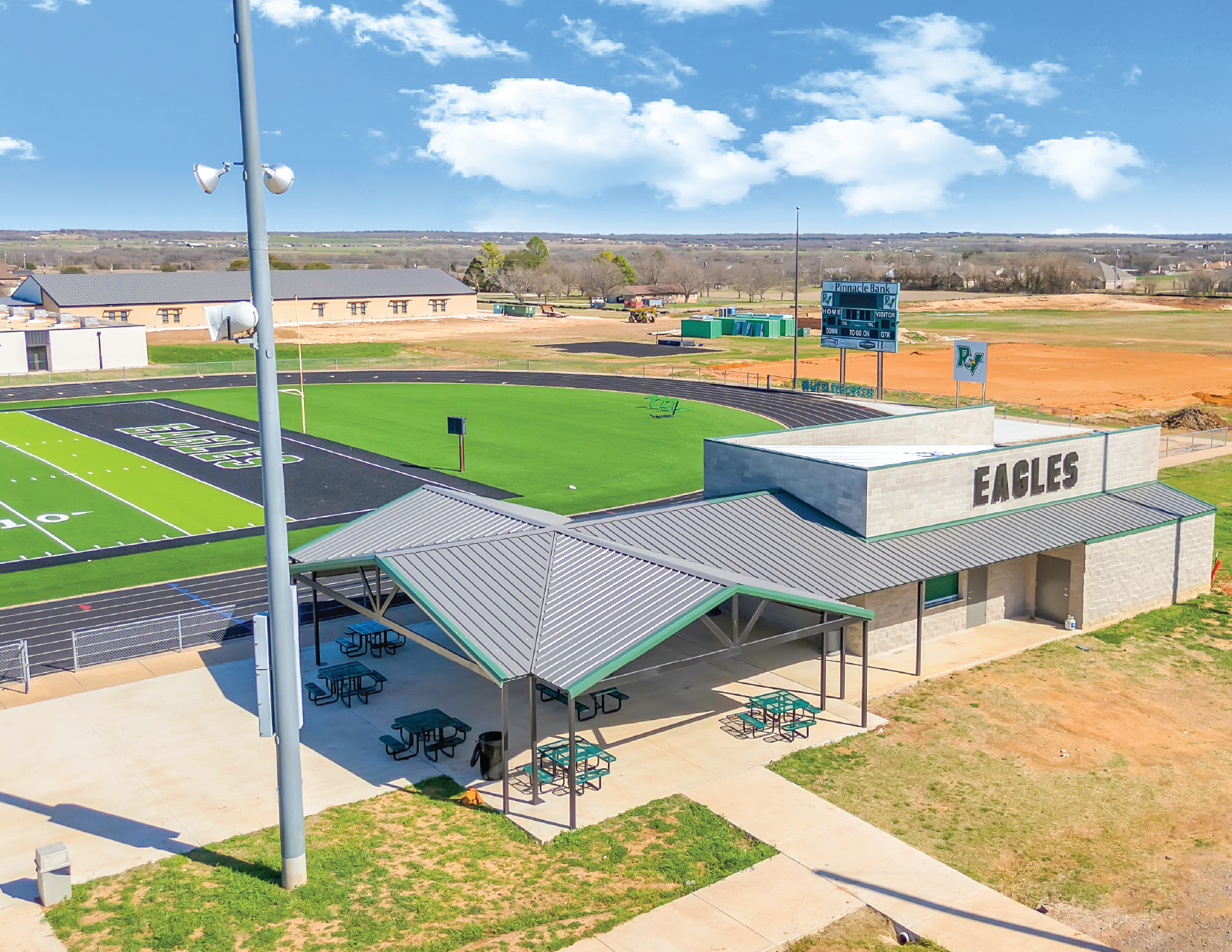 A high school football field with a synthetic turf surface marked with yard lines, a scoreboard, and a large classroom building with the word 