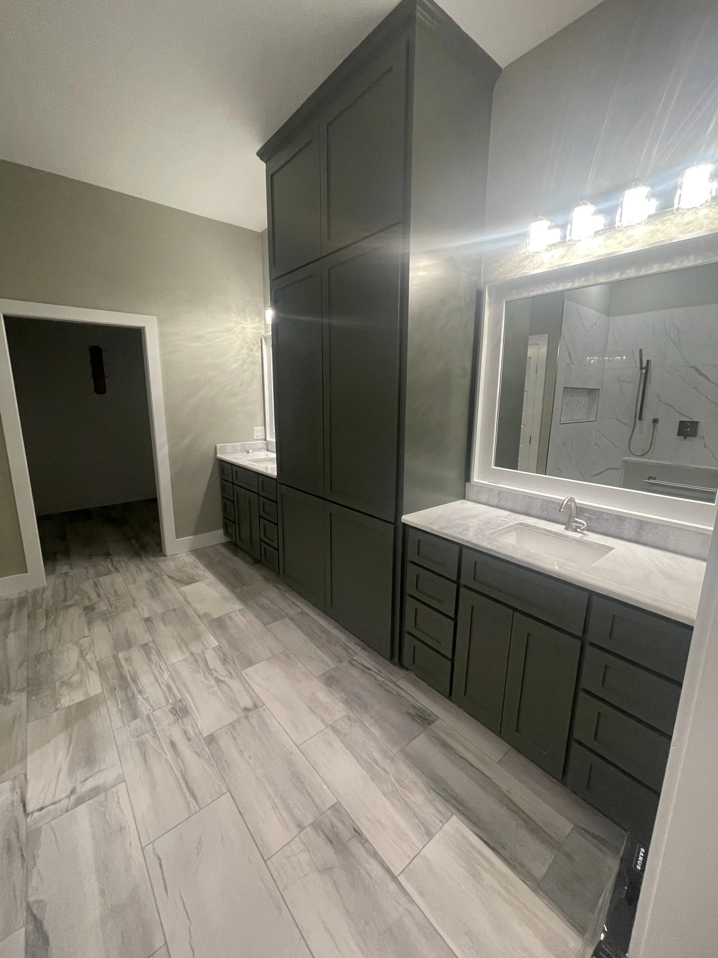 A modern bathroom with gray cabinets, a large mirror, and a marble countertop. The bathroom has a tiled floor and a spacious shower area with marble walls, a shower head, and a built-in shelf.