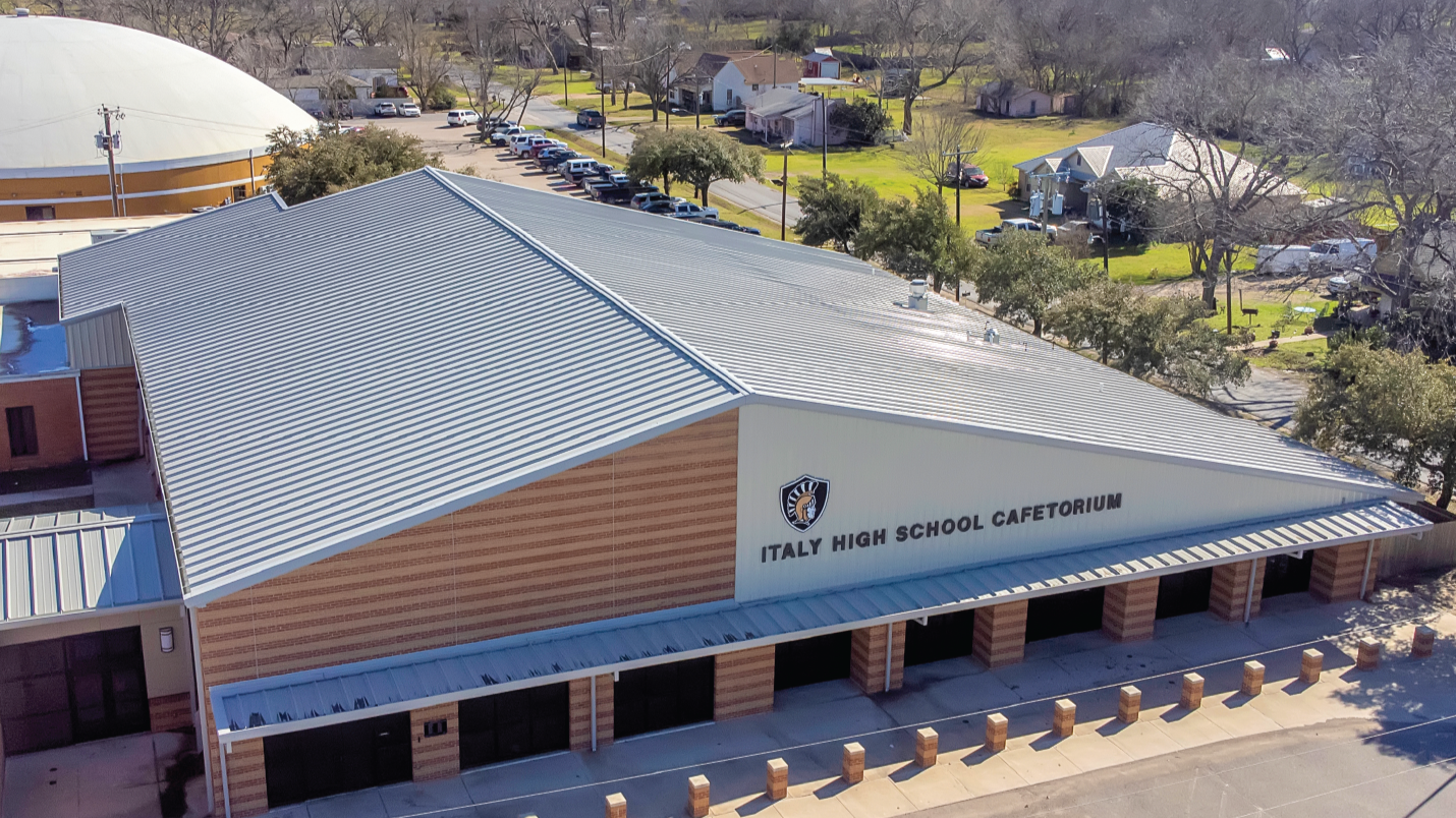 Aerial view of Italy High School Cafetorium with a metal roof and parking lot in front, surrounded by trees and residential houses.